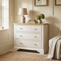 The Amberley 3 Chest of Drawers in Warm White provides compact storage beside a window and a cozy chair, topped with a lamp, books, and flowers for added style.