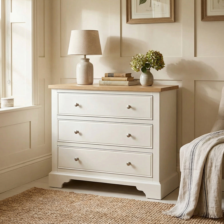 The Amberley 3 Chest of Drawers in Warm White provides compact storage beside a window and a cozy chair, topped with a lamp, books, and flowers for added style.