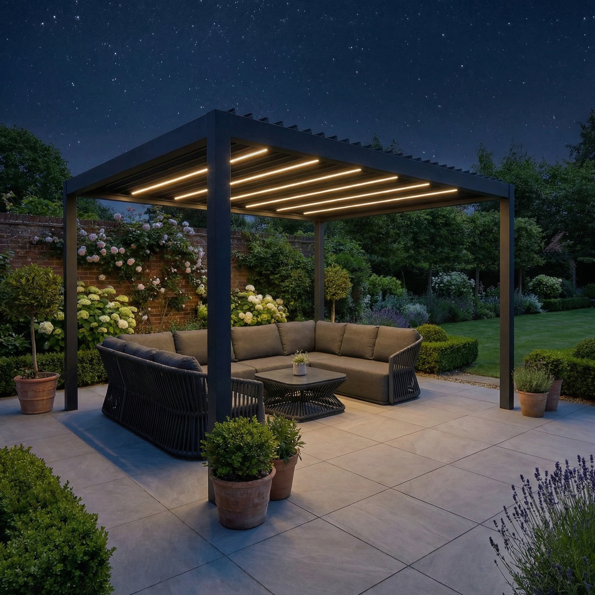 The PergoSTET Deluxe Frame Only 3m x 4m Rectangular Pergola with Motorised LED Louvres in Grey casts a modern glow over a sectional sofa, enhancing a landscaped garden setting at night.