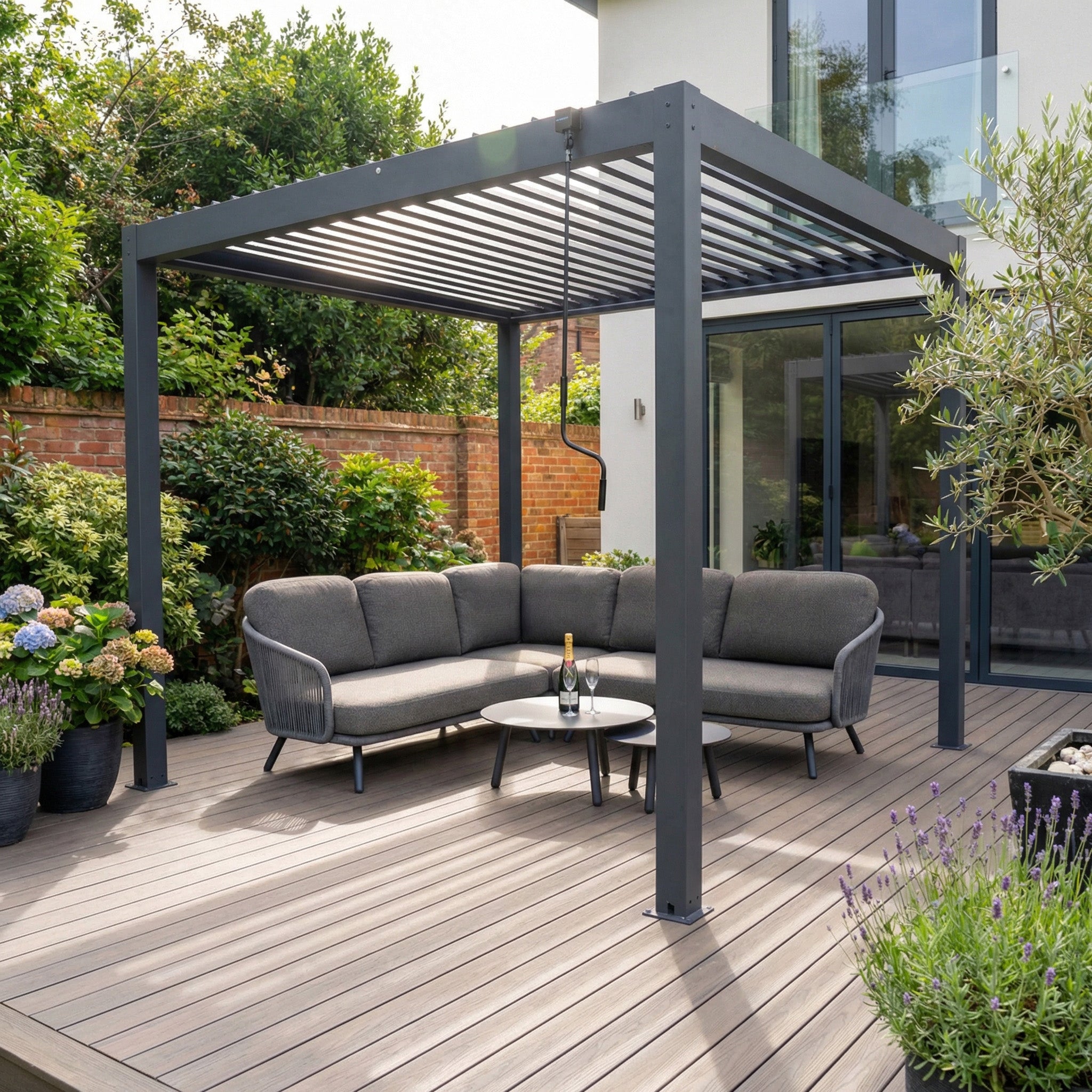Modern patio featuring the PergoSTET Plus 3m x 3m grey pergola with LED lighting, a sectional sofa, round table, and lush plants on a wooden deck.