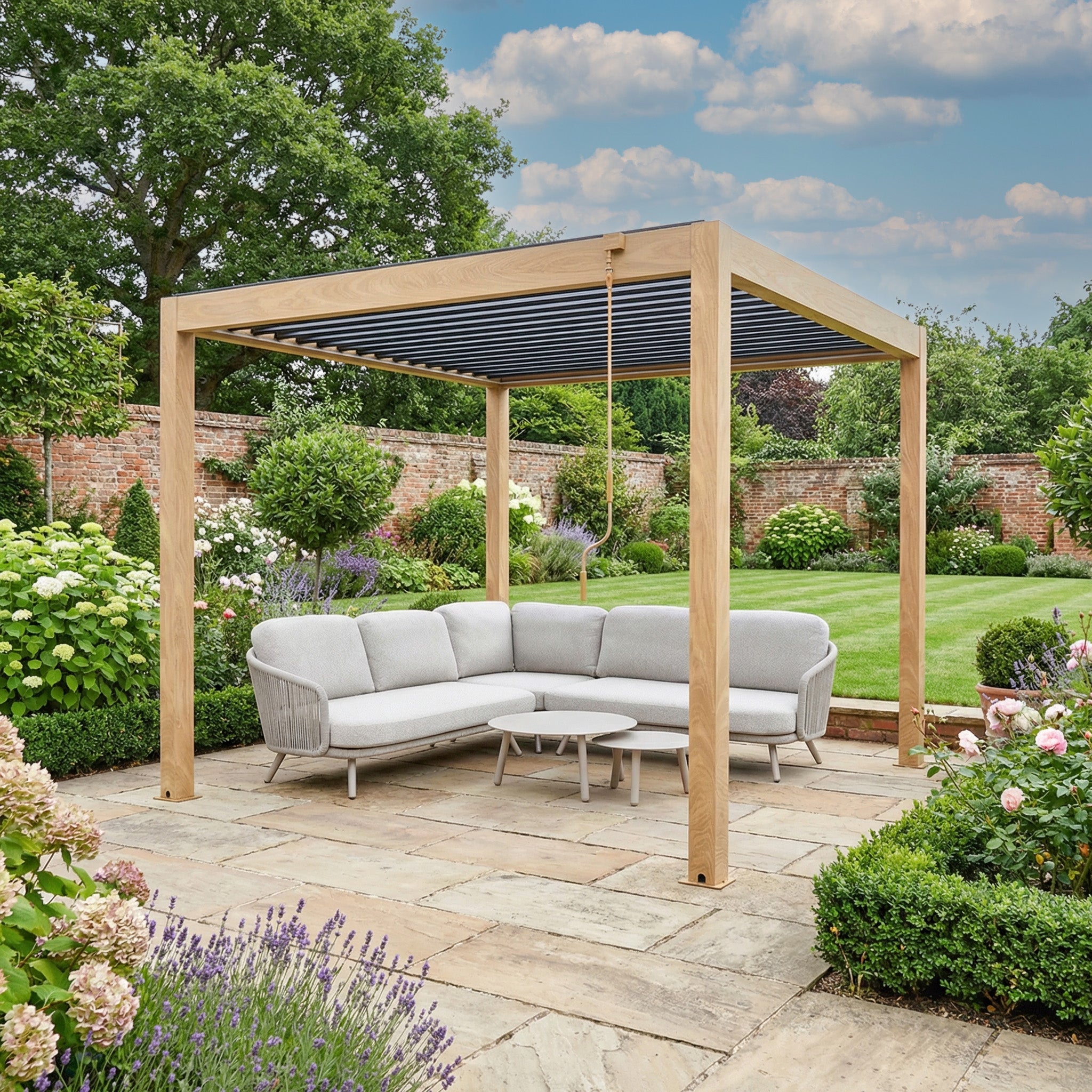The PergoSTET Plus Frame Only 3m x 3m Square Pergola with LED Lighting in Wood Effect creates a stylish outdoor space, paired with a gray sectional sofa and coffee table on a stone patio surrounded by lush garden views.