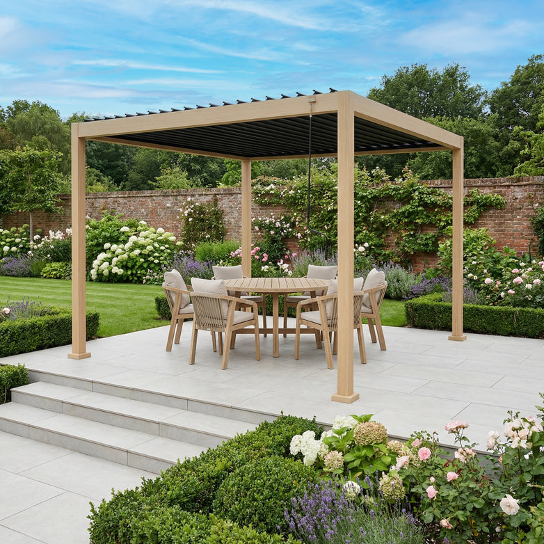 The PergoSTET Lite Neo 3m x 3m Metal Square Pergola in Wood Effect shelters a dining set on a patio, surrounded by flowers and greenery in a beautifully landscaped garden.