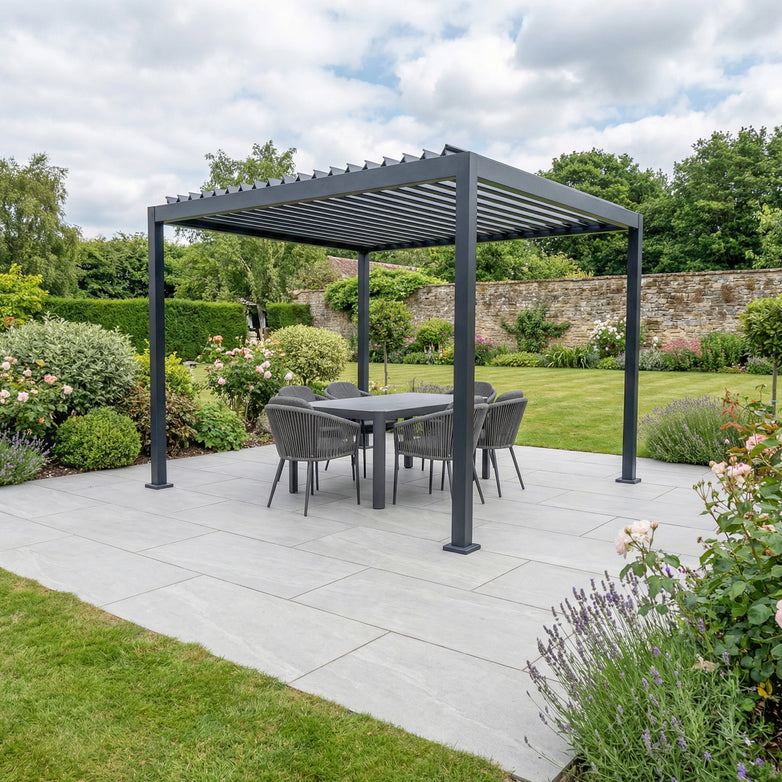 The PergoSTET Lite Neo 3m x 3m Metal Square Pergola in Grey with tilting louvres and built-in drainage shelters a dining set on a tiled patio amid a lush flower and shrub garden.