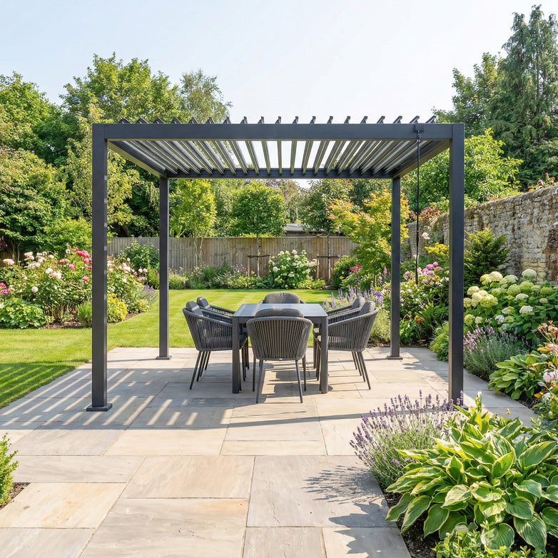 The PergoSTET Lite Neo 3m x 3m Metal Square Pergola in Grey, with tilting louvres, is featured alongside a dining table and chairs in a landscaped garden with lush plants and a stone patio.