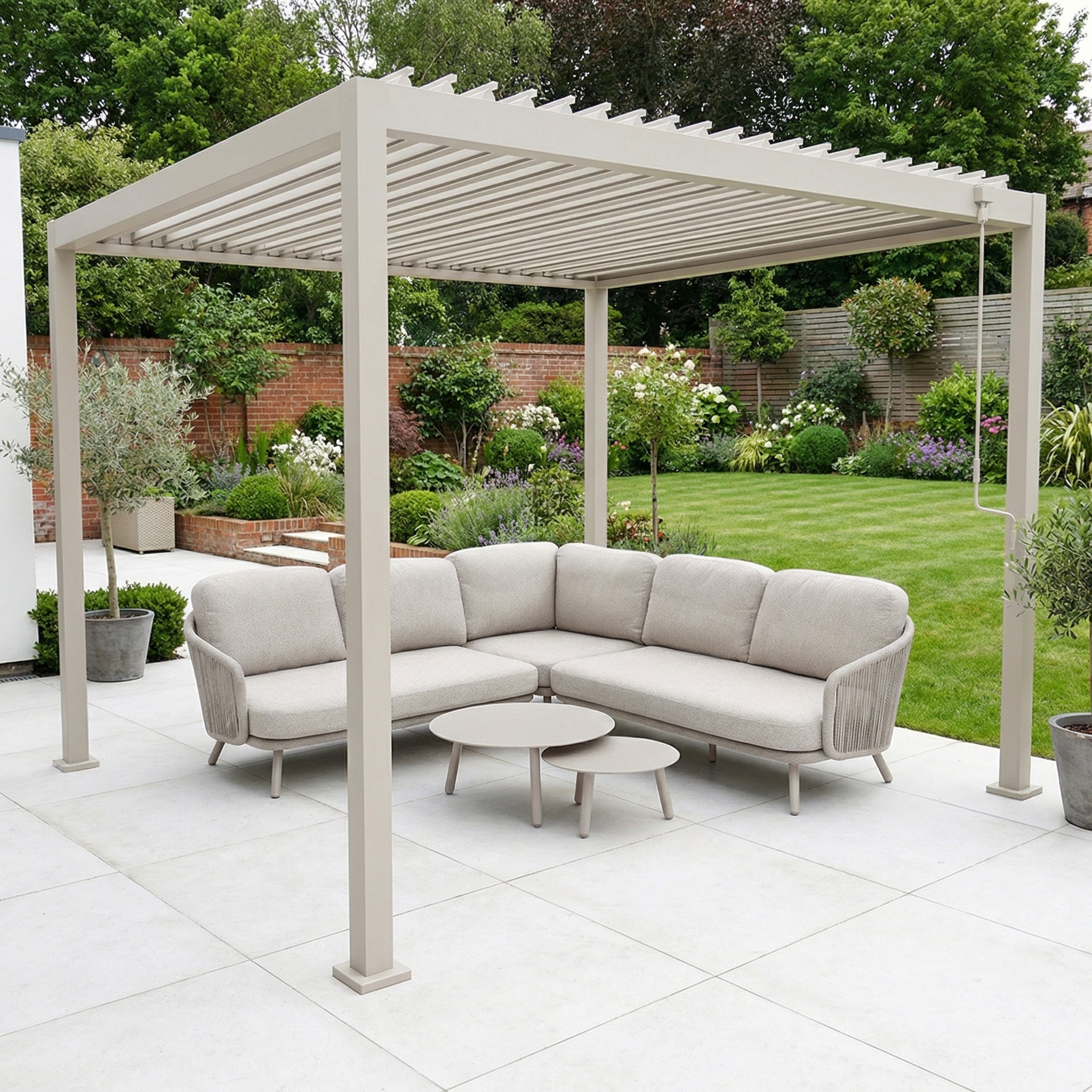 The PergoSTET Lite 3m x 3m Metal Square Pergola in Latte is paired with a beige outdoor sofa and round tables on a patio, all set within a lush green garden.
