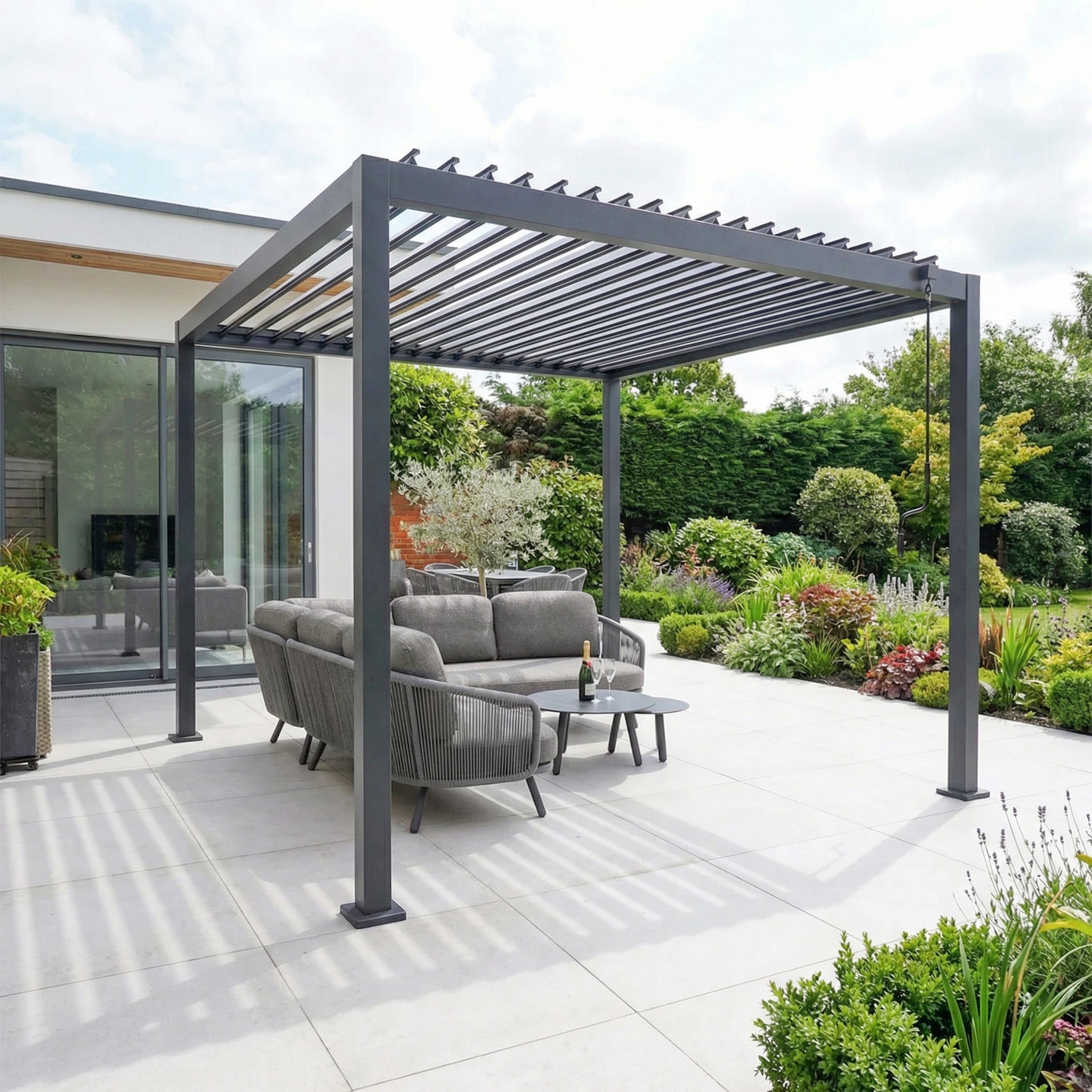 The PergoSTET Lite 3m x 3m Metal Square Pergola in Grey features tilting louvres and outdoor furniture on a patio, set beside a lush garden.