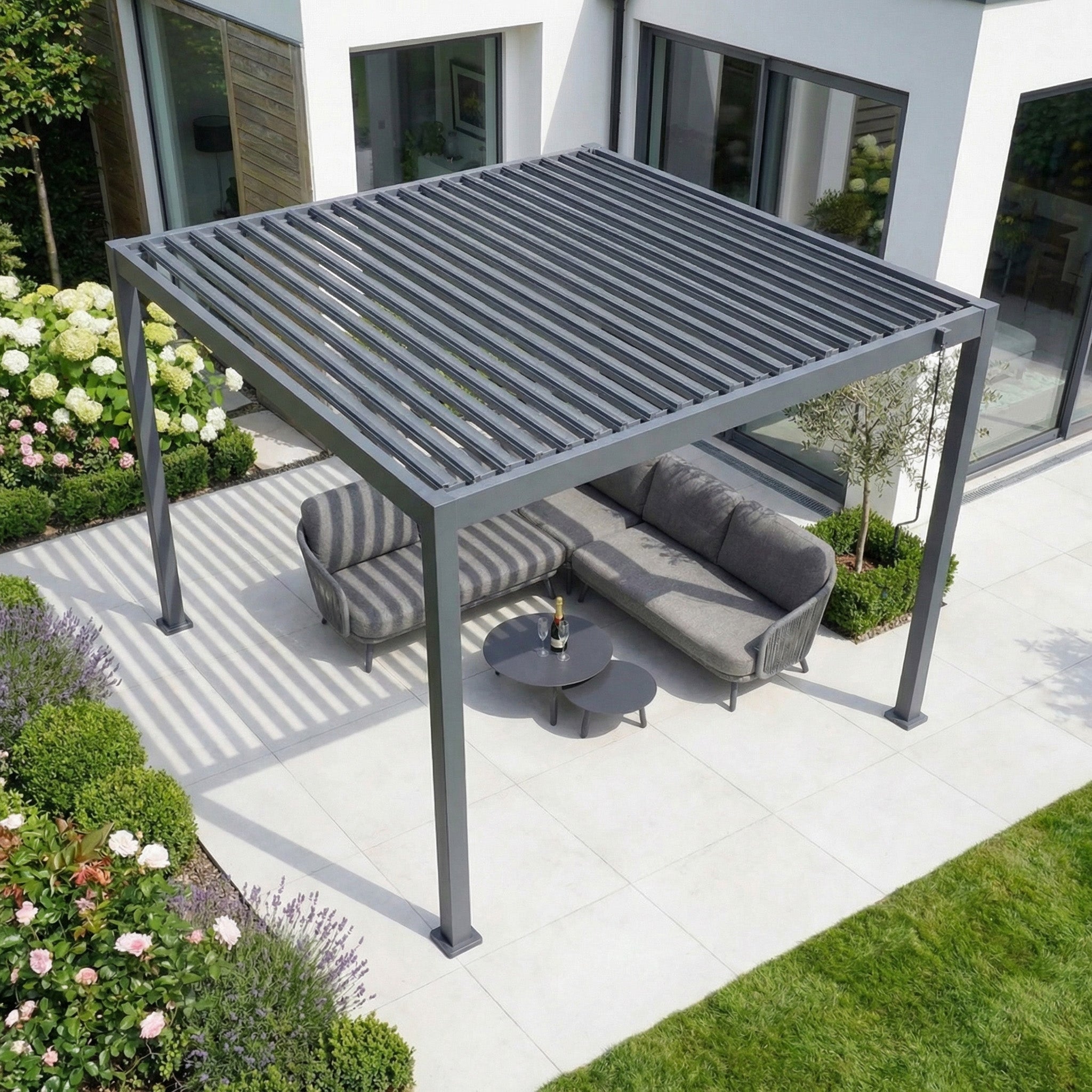 Contemporary patio with the PergoSTET Lite 3m x 3m Metal Square Pergola in Grey, tilting louvres, cushioned seating, round table, and lush garden landscaping.
