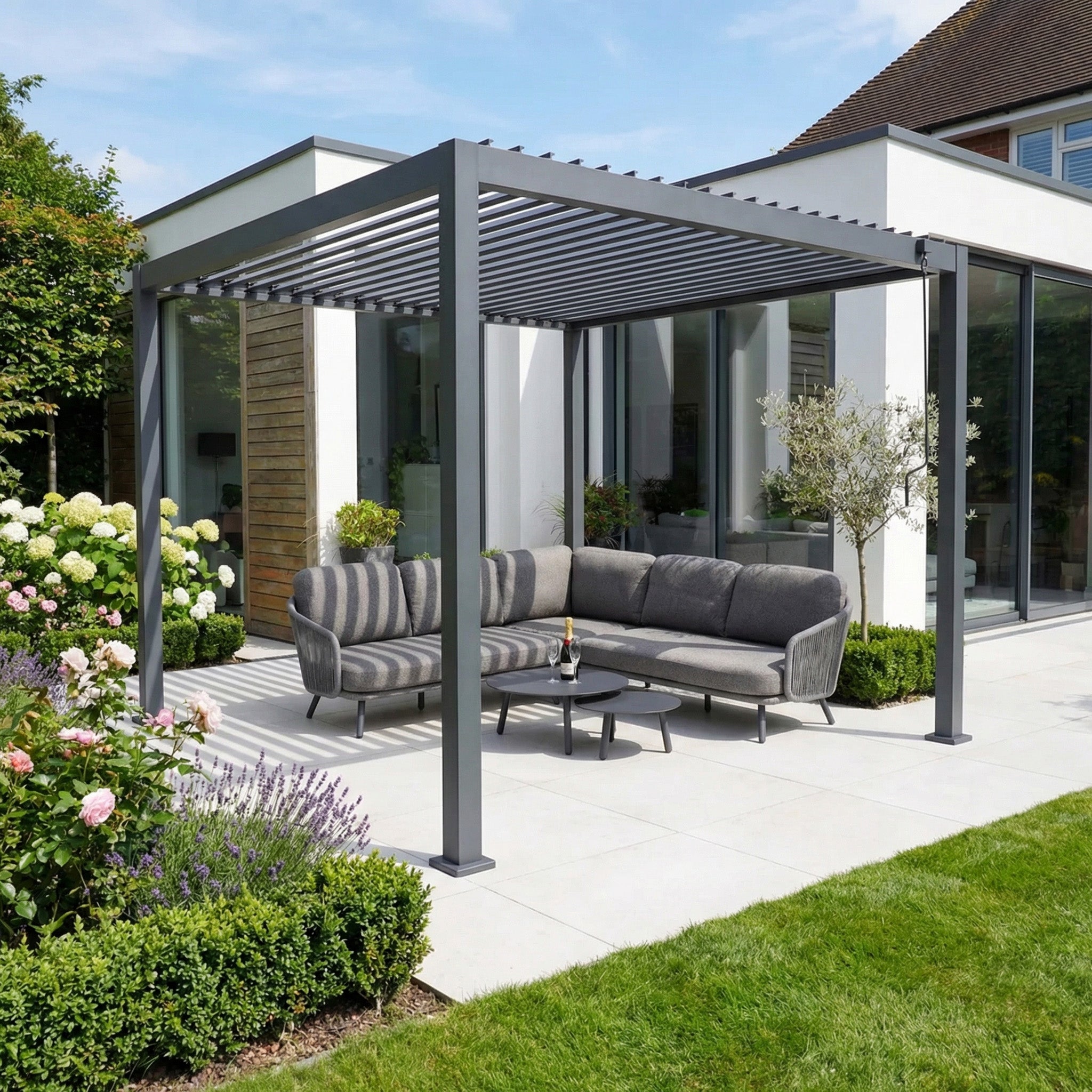 Modern patio with a PergoSTET Lite 3m x 3m Metal Square Pergola in Grey featuring tilting louvres, gray outdoor sofa, small table, and lush garden in front of a white house.