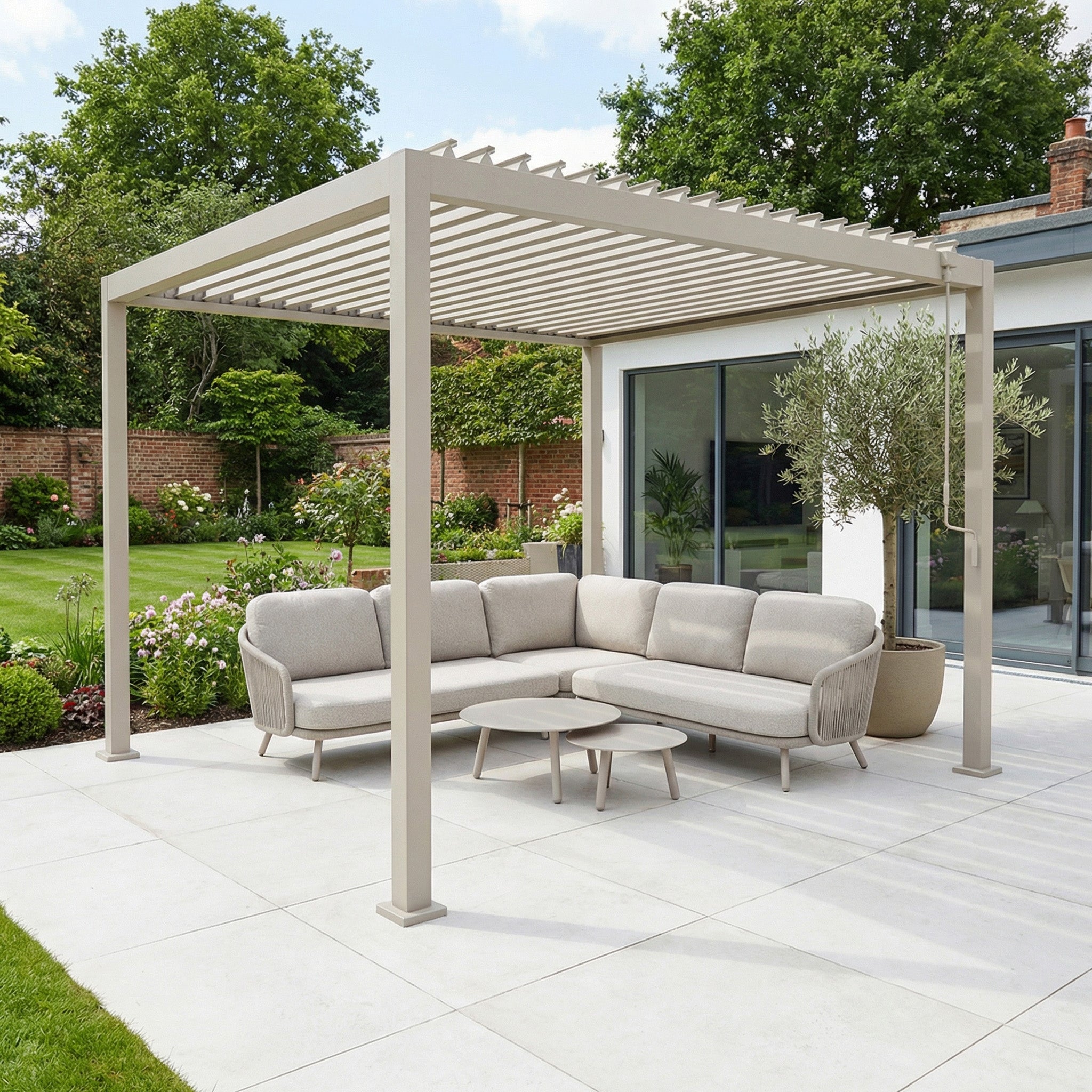 The PergoSTET Lite 3m x 3m Metal Square Pergola in Latte frames a beige outdoor sofa and round table on a patio, set within a landscaped garden.