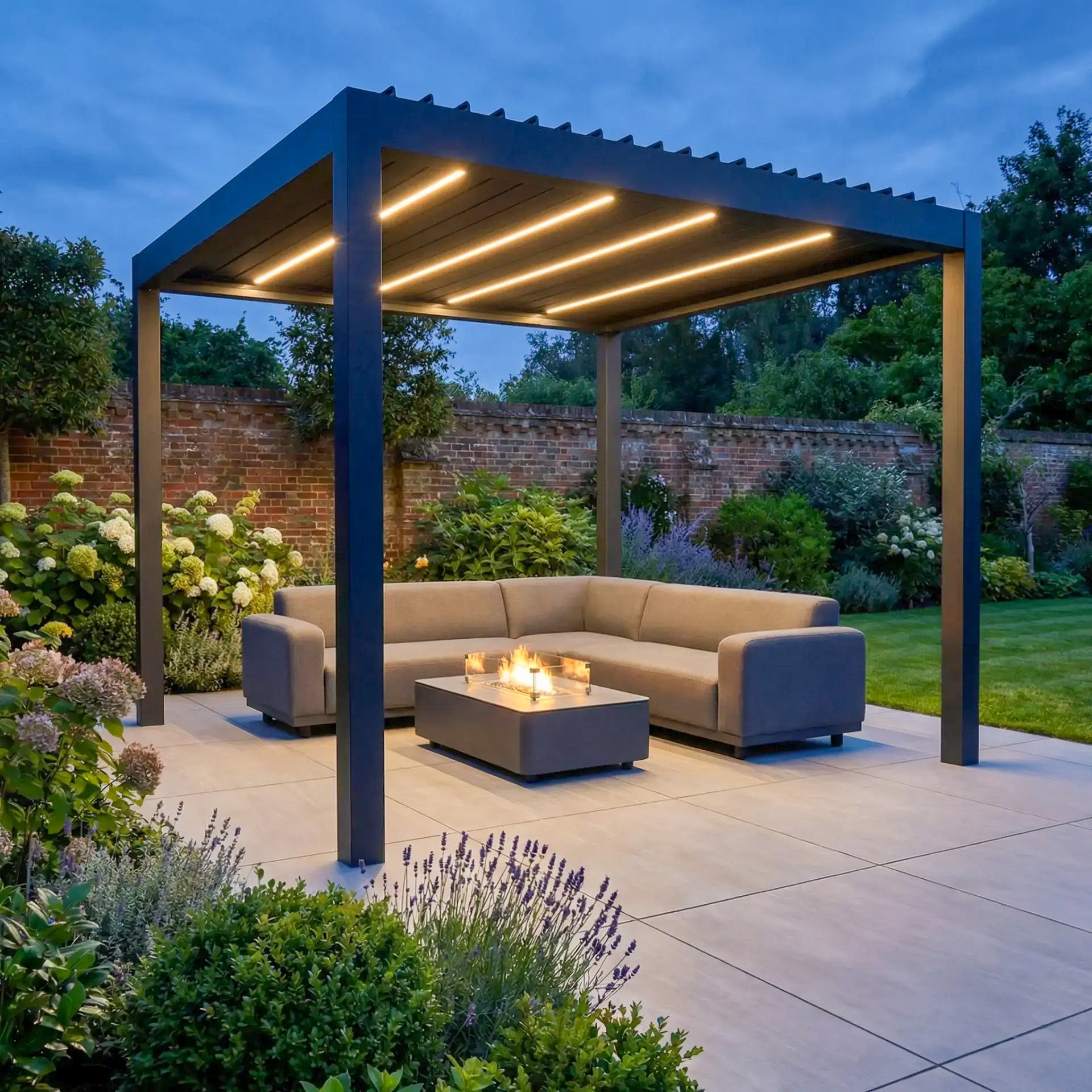 Modern outdoor patio with a PergoSTET Deluxe 3m x 3m Square Pergola (motorised LED louvres, grey), gray sectional sofa, and fire pit, surrounded by lush garden plants.