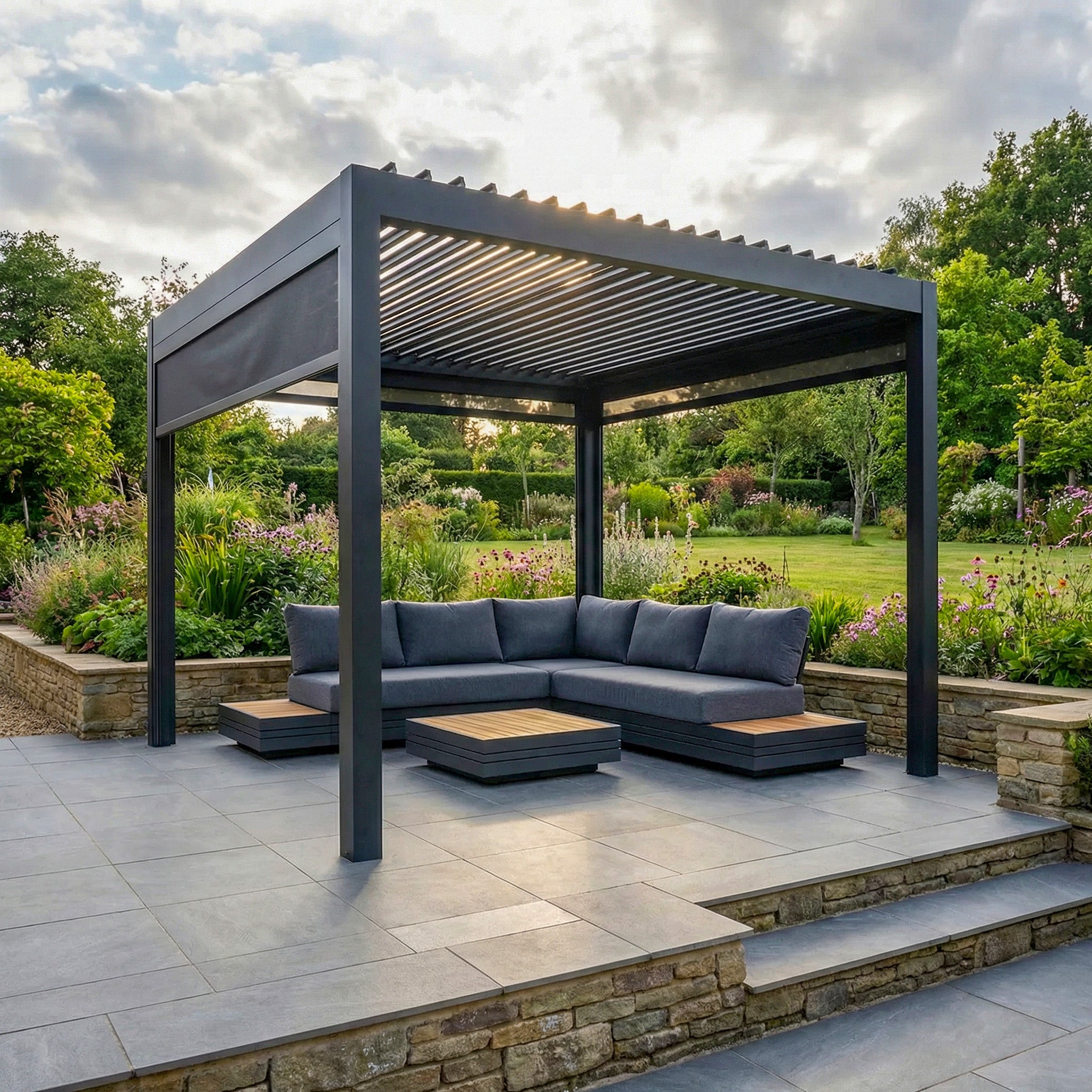 The PergoSTET Deluxe 3m x 3m Square Pergola features motorised LED louvres and 3 manual side blinds in grey, set on a tiled patio with lush greenery, paired with a gray sectional sofa and tables.