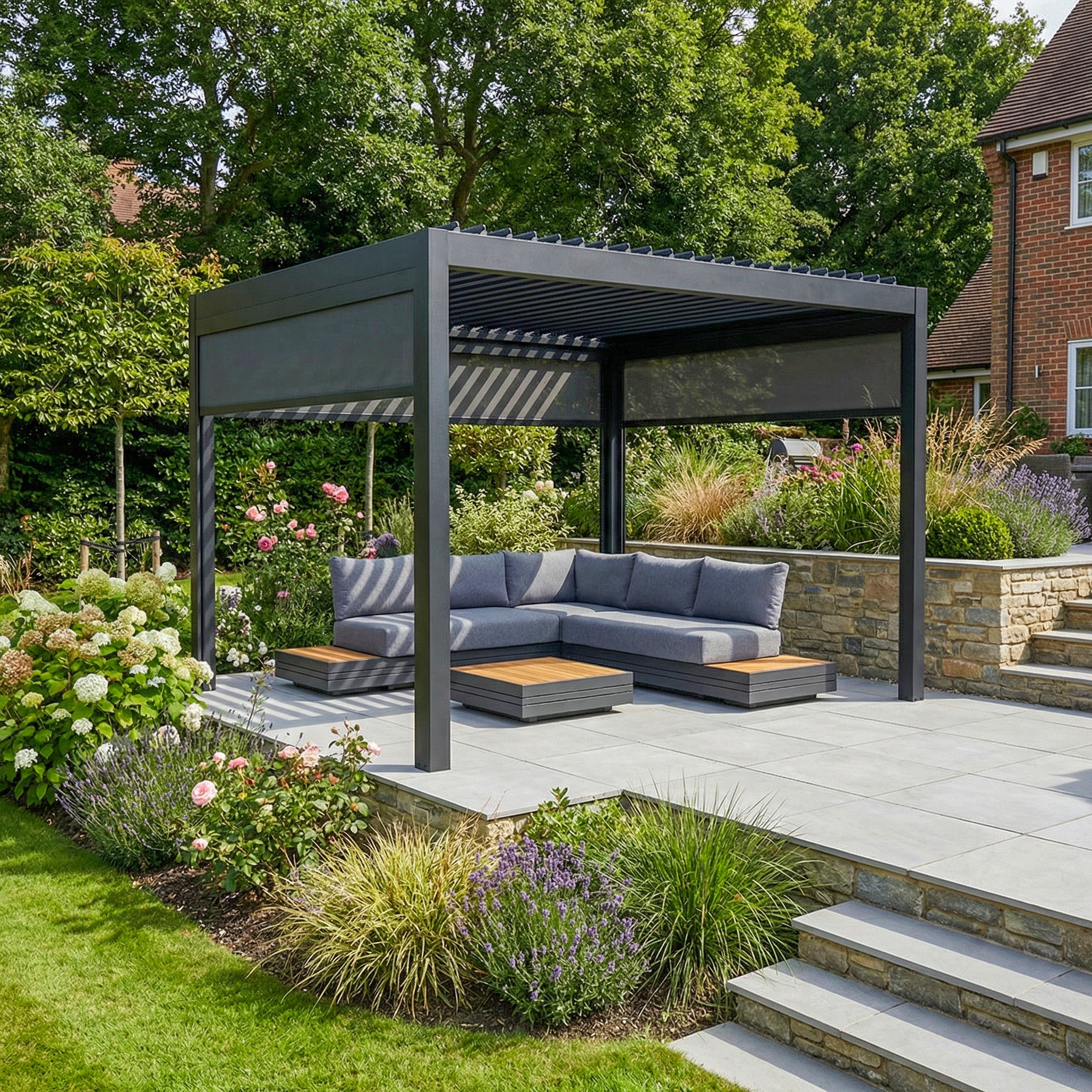The PergoSTET Deluxe 3m x 3m Square Pergola features motorised LED louvres and 3 manual side blinds in grey, paired with stylish outdoor seating on a stone patio amidst lush green garden landscaping.