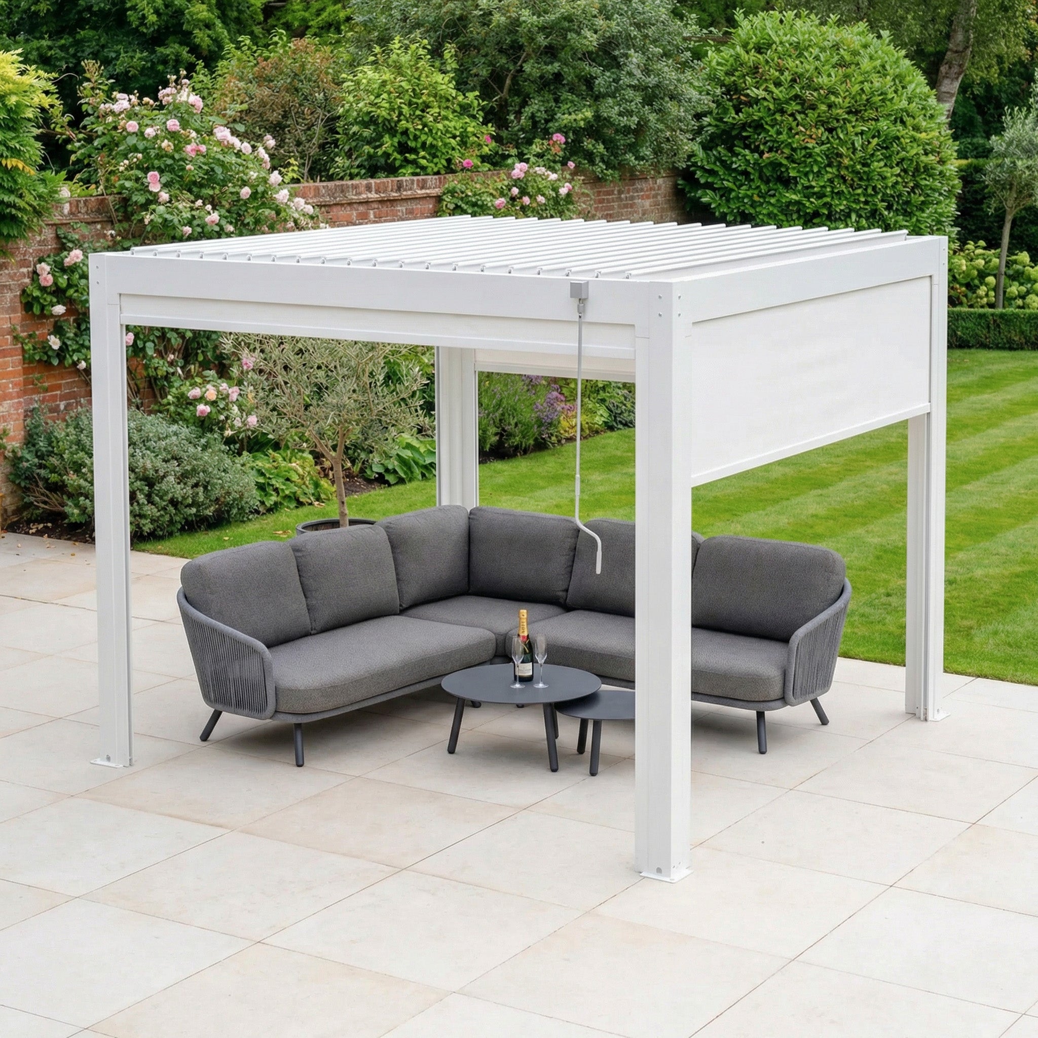 The PergoSTET Plus 3m x 3m White Pergola with manual side blinds and LED lighting shelters a gray outdoor sofa set and round table on a tiled patio, surrounded by a lush green garden.