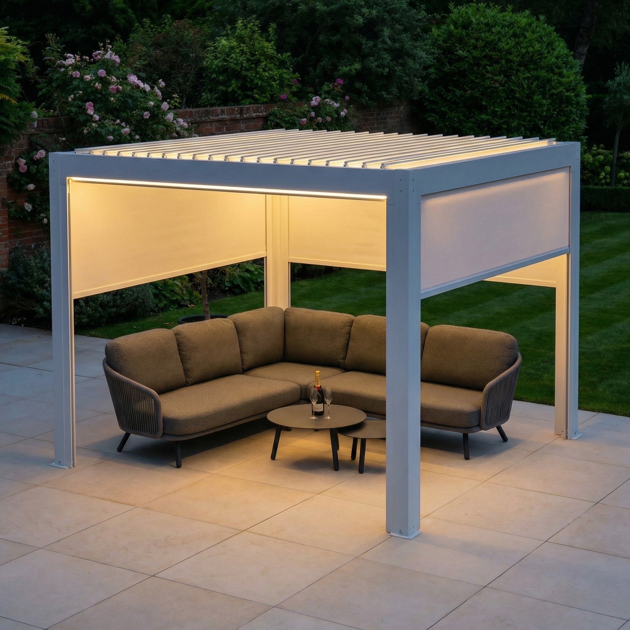 The PergoSTET Plus 3m x 3m Square Pergola in white features 3 manual side blinds and LED lighting, providing cozy outdoor seating on the patio and blending beautifully with a lush green garden.