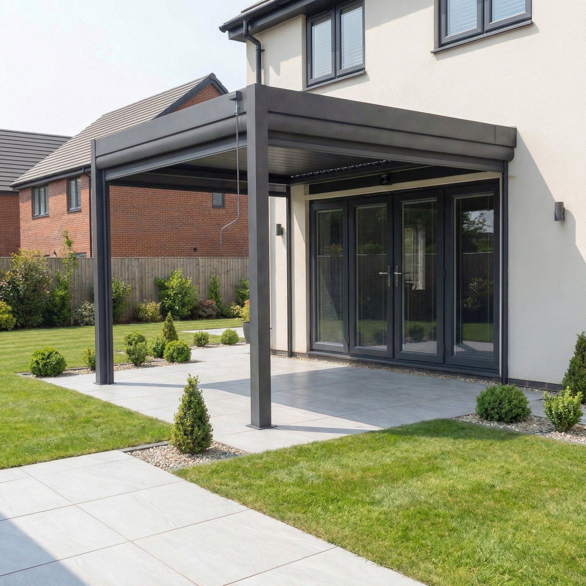 A modern patio with a grey PergoSTET Plus 3m x 3m square wall mounted pergola, featuring tilting louvres, three manual side blinds, and LED lighting, attached to the house and surrounded by lawn and shrubs.