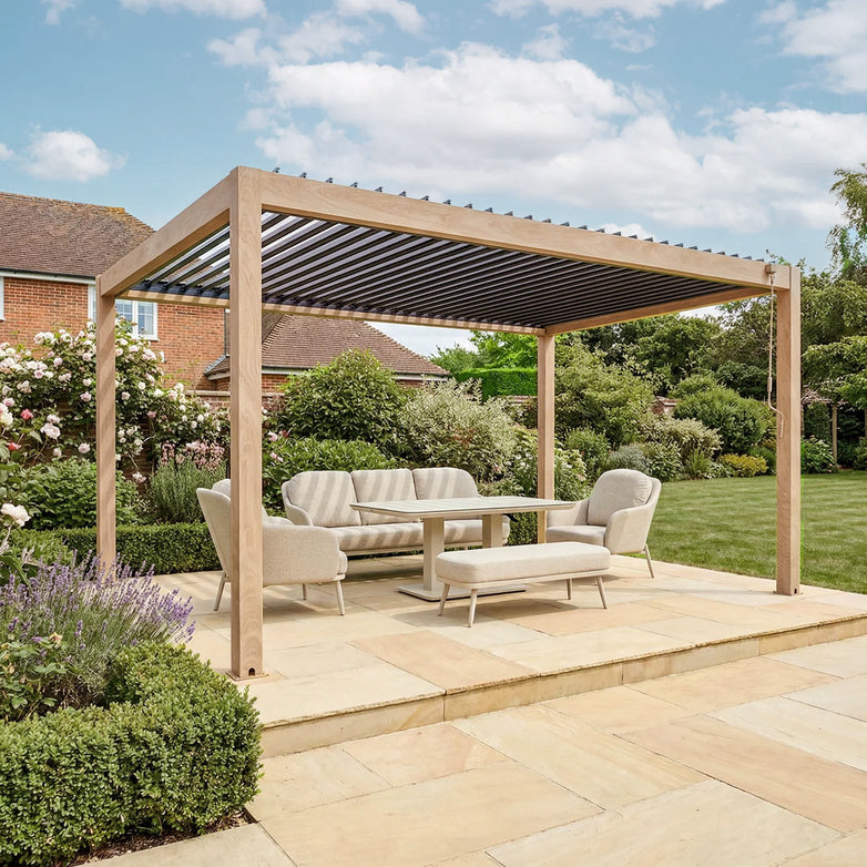 The PergoSTET Plus Frame Only 3m x 4m rectangular pergola with LED lighting in wood effect stands on a stone patio, providing stylish shelter in a lush garden filled with flowers and shrubs.