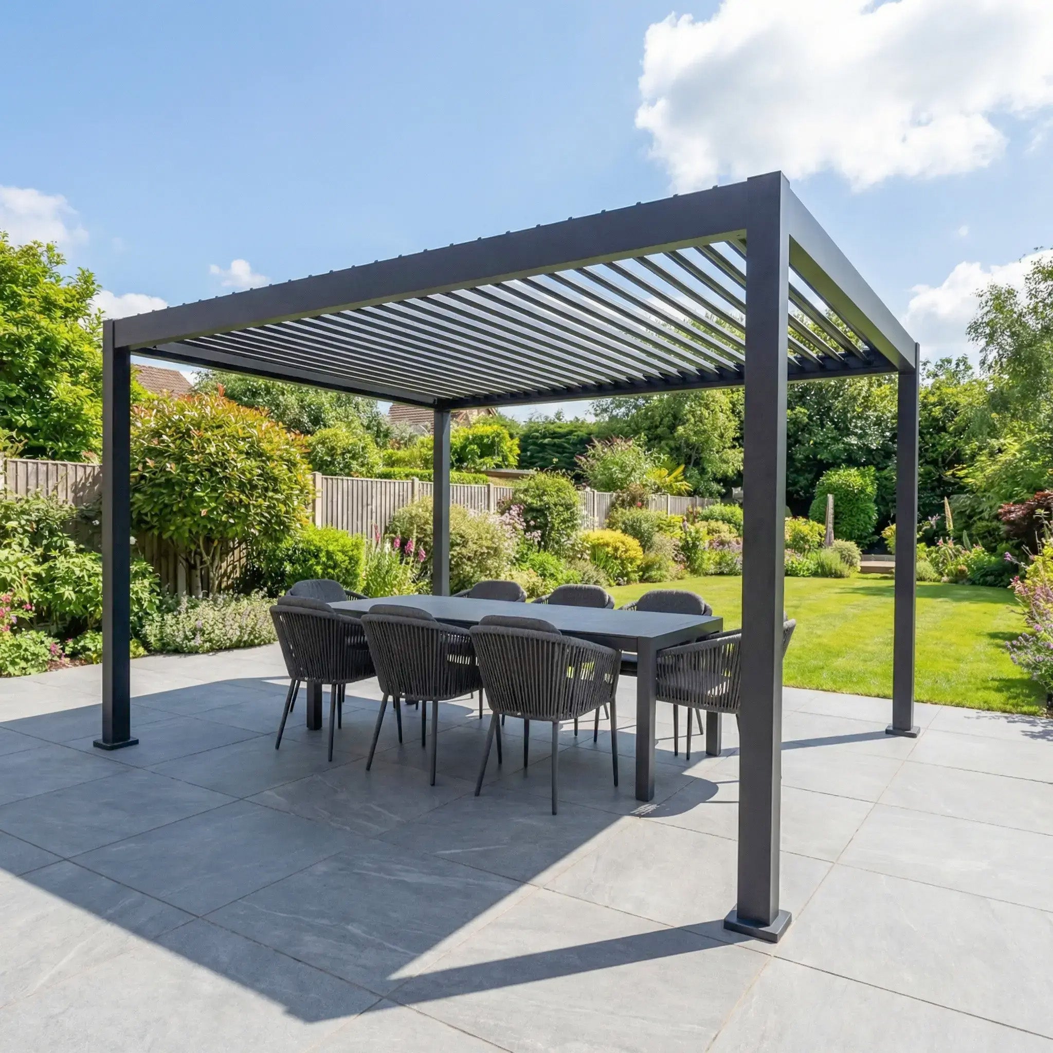 The PergoSTET Lite Neo 3m x 4m Metal Rectangular Pergola in Grey features a louvred roof, built-in drainage, and is set on a patio with a dining area, surrounded by greenery under a clear sky.