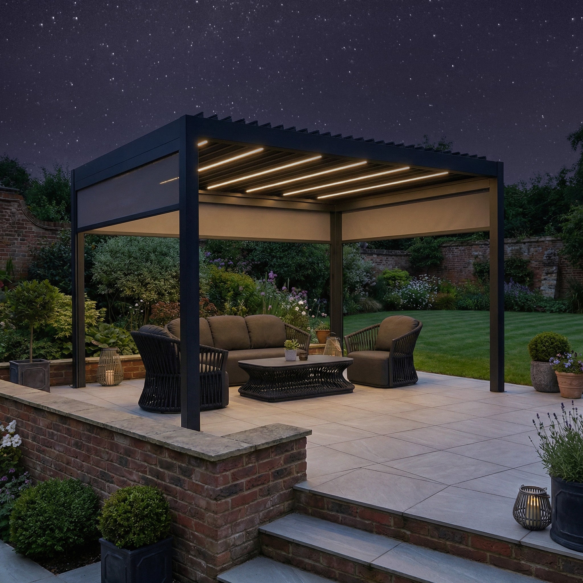A garden seating area at night beneath the PergoSTET Deluxe 3m x 4m Rectangular Pergola, featuring grey motorised LED louvres and three manual side blinds, illuminated by string lights under a starry sky.