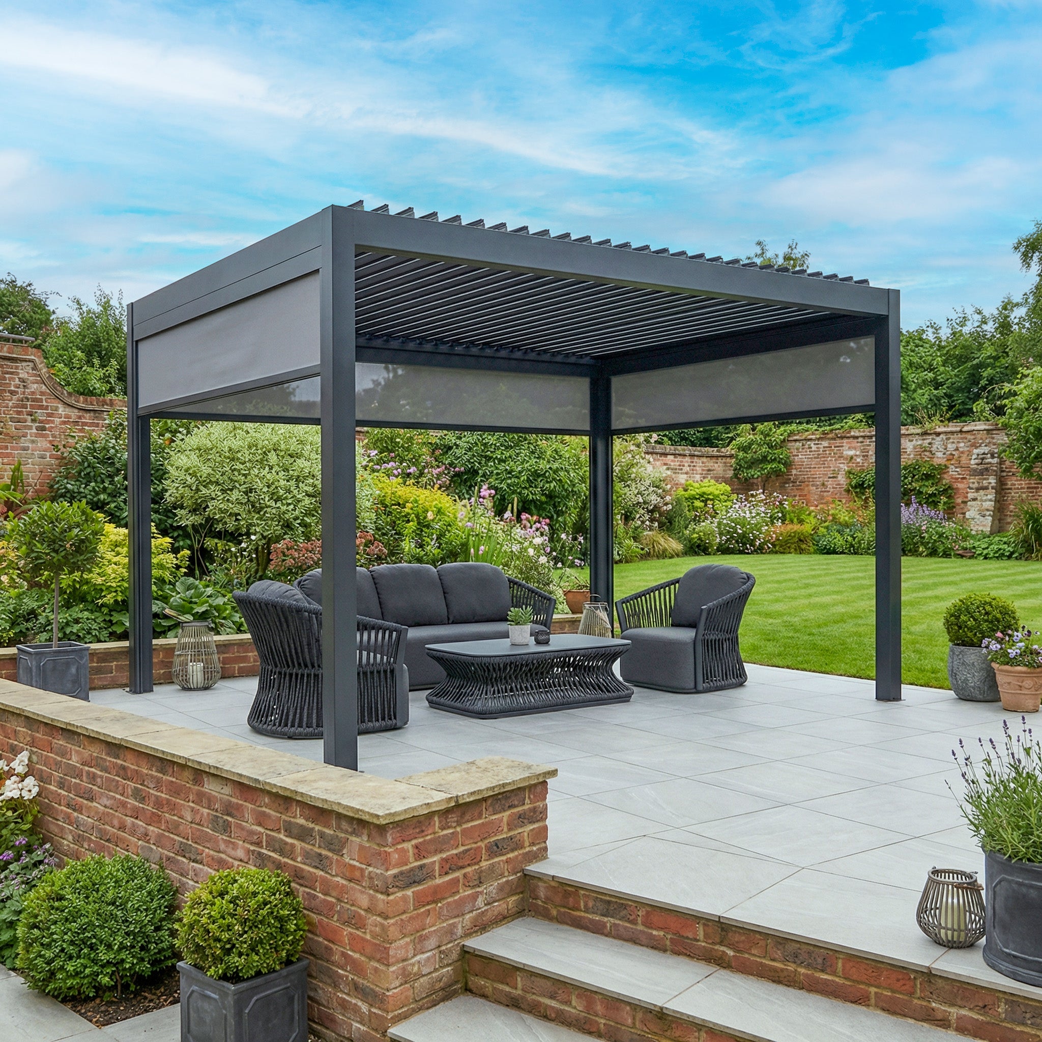 Enhance your patio with the PergoSTET Deluxe 3m x 4m Rectangular Pergola in grey, featuring motorised LED louvres and 3 manual side blinds—ideal for stylish outdoor living amid your garden greenery.