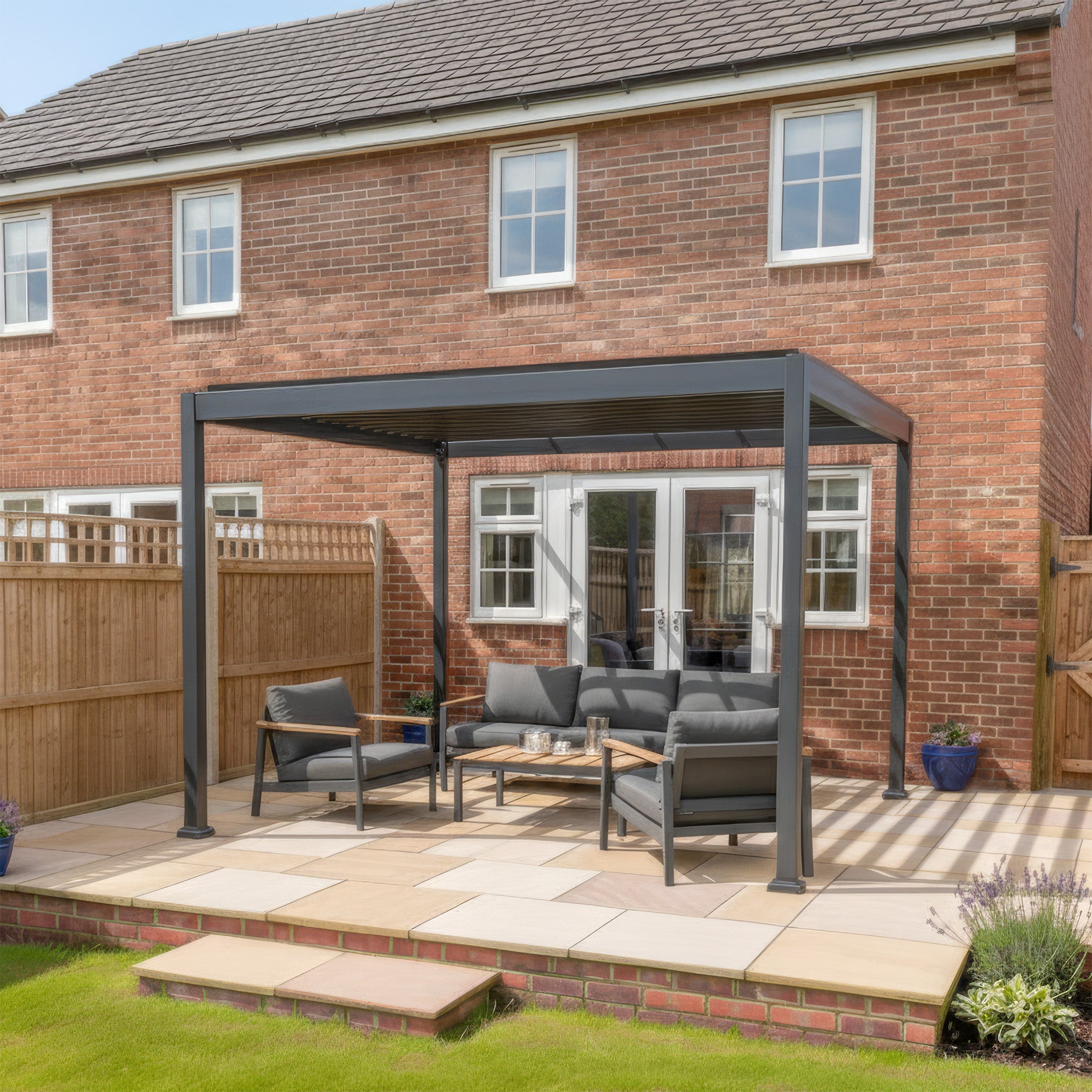 The PergoSTET Lite Neo 3m x 4m Metal Rectangular Pergola in Grey adds a modern touch with tilting louvres and outdoor seating on a stone patio in the backyard of a brick house.