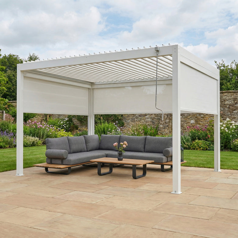 The PergoSTET Plus 3m x 4m rectangular pergola with 3 manual side blinds and LED lighting in white sits on a stone patio with drainage, featuring a gray sofa set and coffee table, all surrounded by lush greenery.