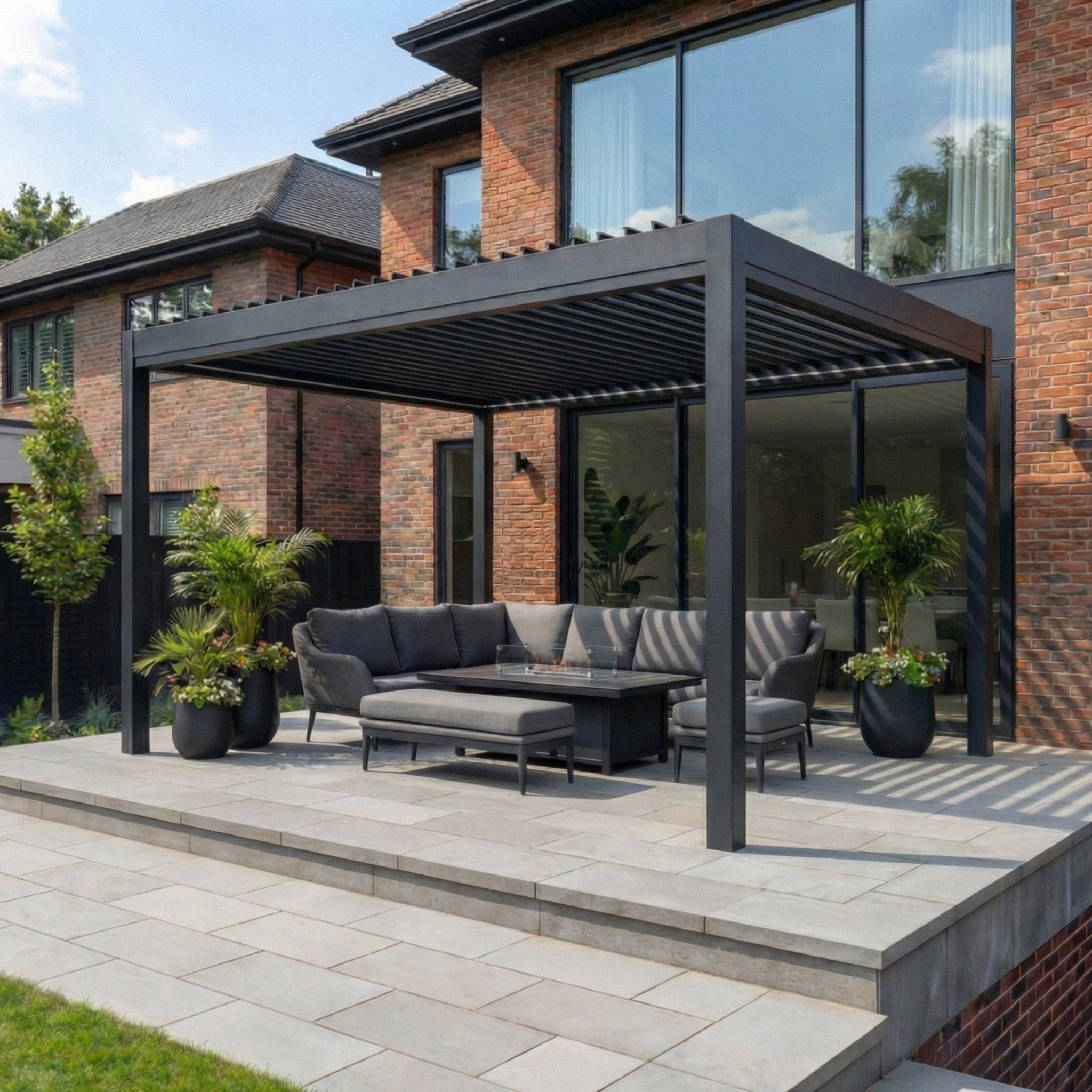 A modern patio features the PergoSTET Pro Frame Only 3m x 4m Rectangular Pergola with LED Lighting in Grey, set in front of a brick house and surrounded by plants, complemented by gray outdoor furniture.