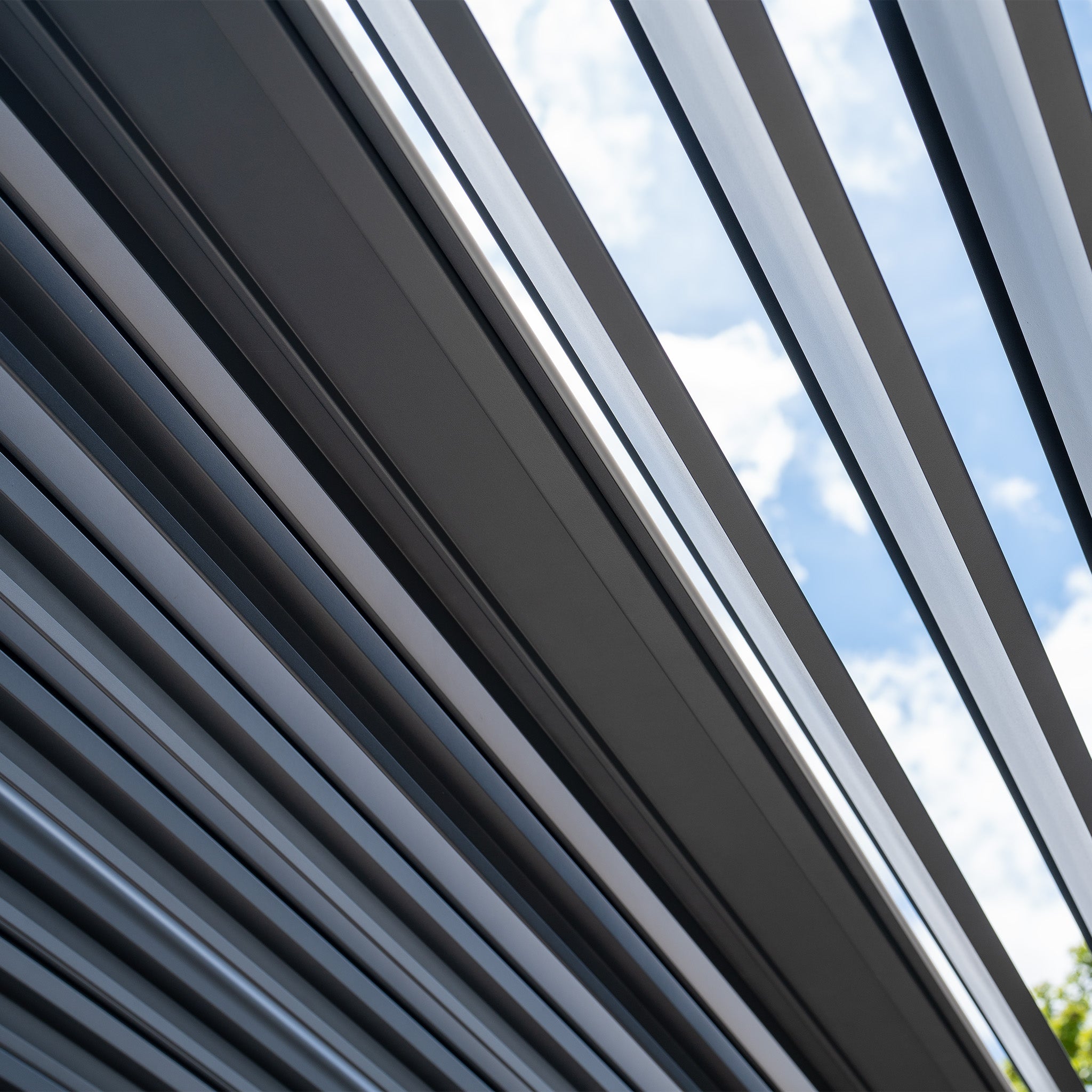 The PergoSTET Deluxe 3m x 6m Rectangular Pergola, featuring motorised LED louvres and four manual side blinds in grey, effortlessly blends style and functionality to create captivating light and shadow patterns against a partly cloudy sky.
