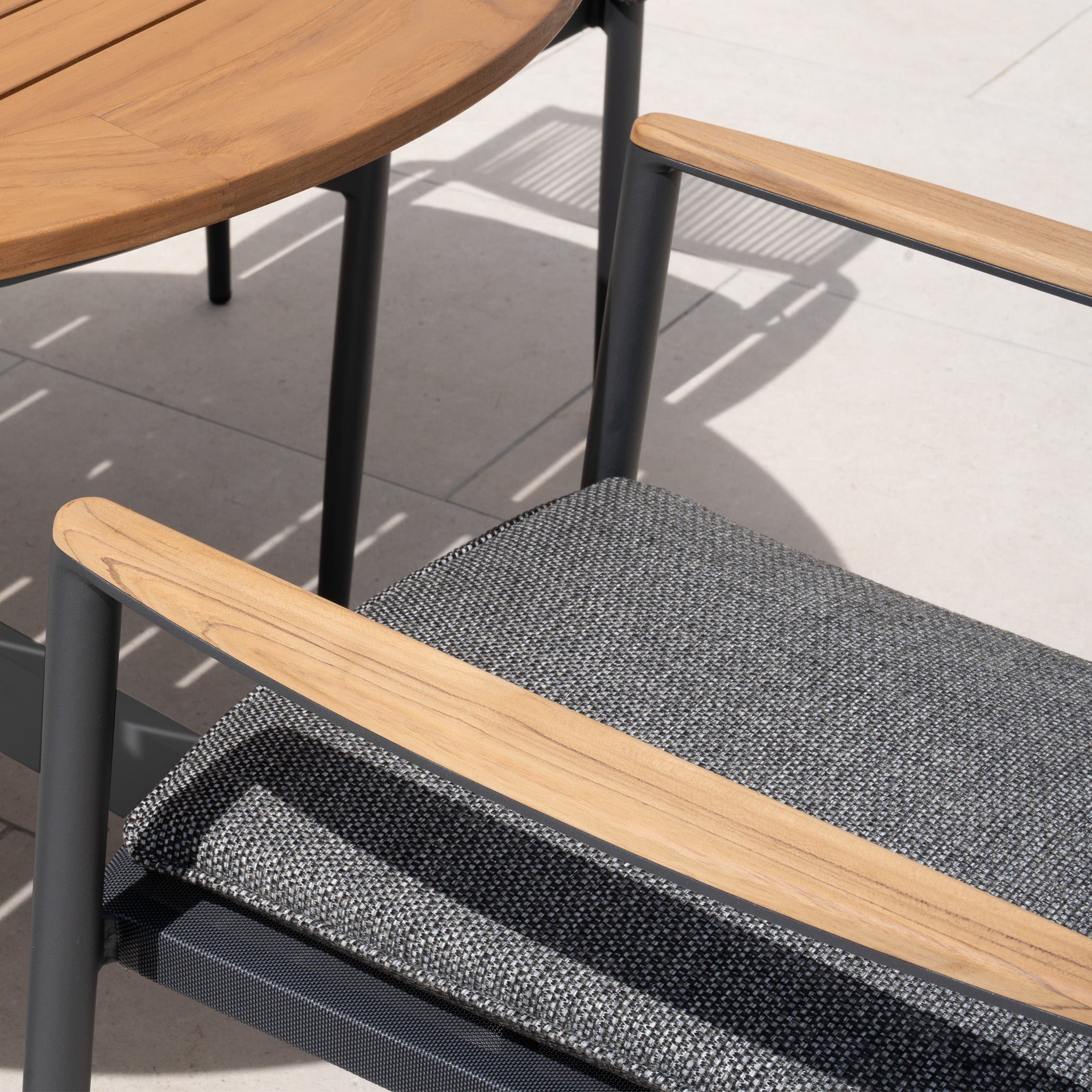 Close-up of Tellaro Rope Dining Chair in Grey (4pcs) with wooden armrests beside a round wooden table on a light stone patio.