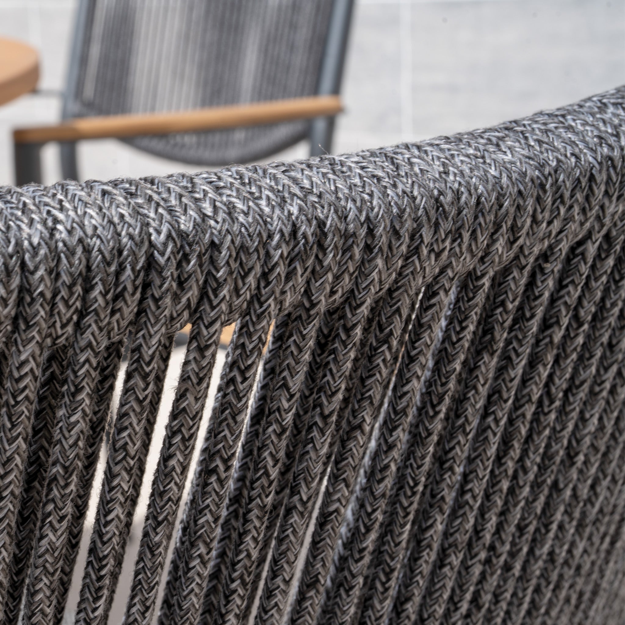 Close-up of a Tellaro Rope Dining Chair in Grey (4pcs) featuring a gray woven rope back, with another chair and a table softly blurred in the background.