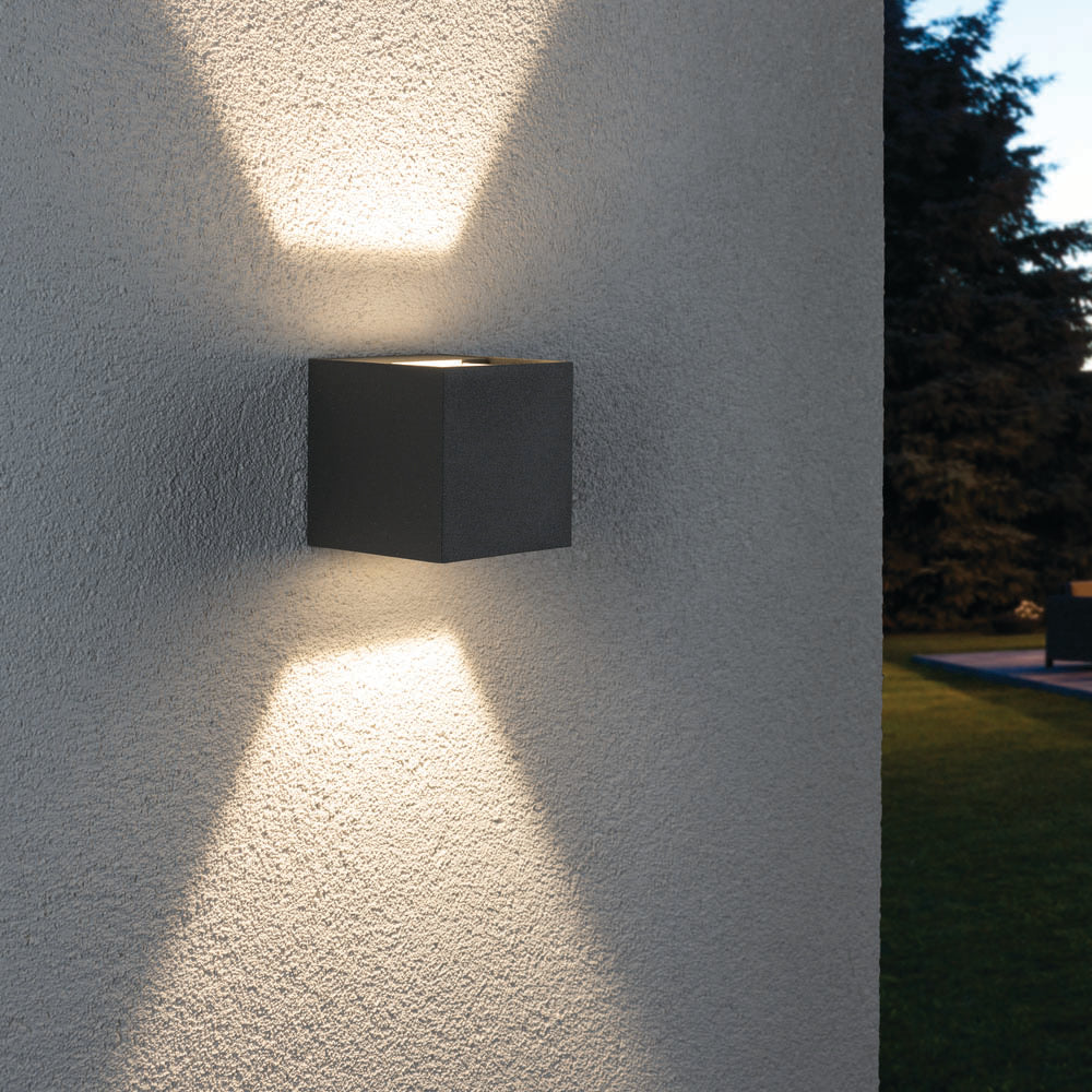 The Paulmann - Outdoor Cybo 7.7W LED 8x8cm Wall Light in Grey creates an X-shaped pattern with captivating light effects on a textured exterior wall, beautifully complementing a yard and trees. Its adjustable beam angle provides versatile lighting options to enhance any outdoor space.
