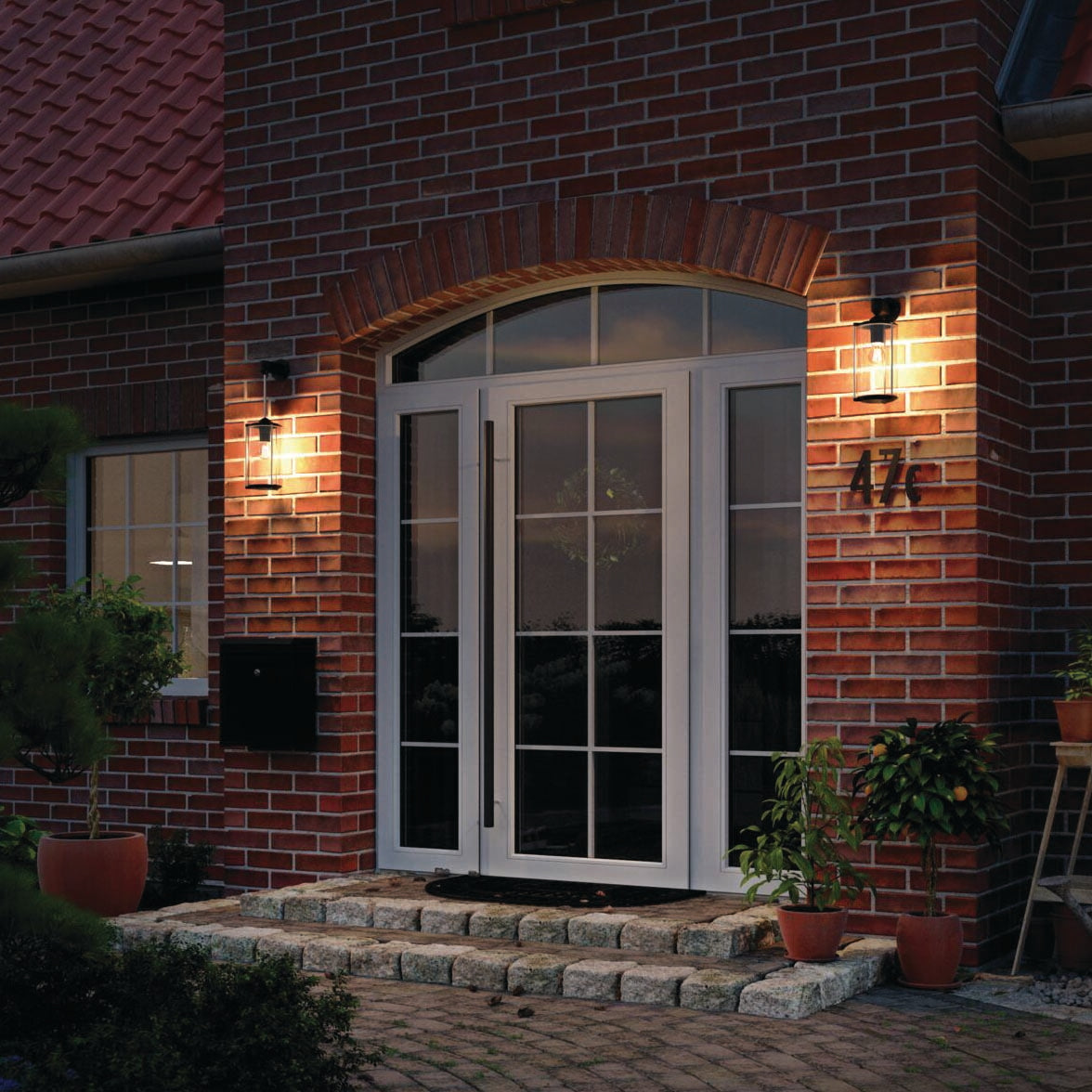 The front entrance of the brick house showcases a white door, warmly lit by two wall-mounted LED lamps with Paulmann Filament LED Pear Bulbs 15W E27 in warm white, while pots of vibrant plants line the steps.