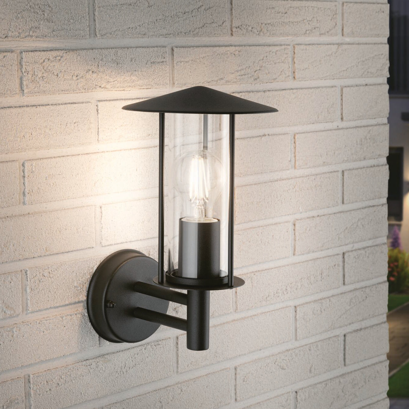 Outdoor wall light fixture on white brick wall, featuring a glass enclosure and black metal frame with an elegant Paulmann Filament LED Pear Bulb 15W E27 Warm White lamp.