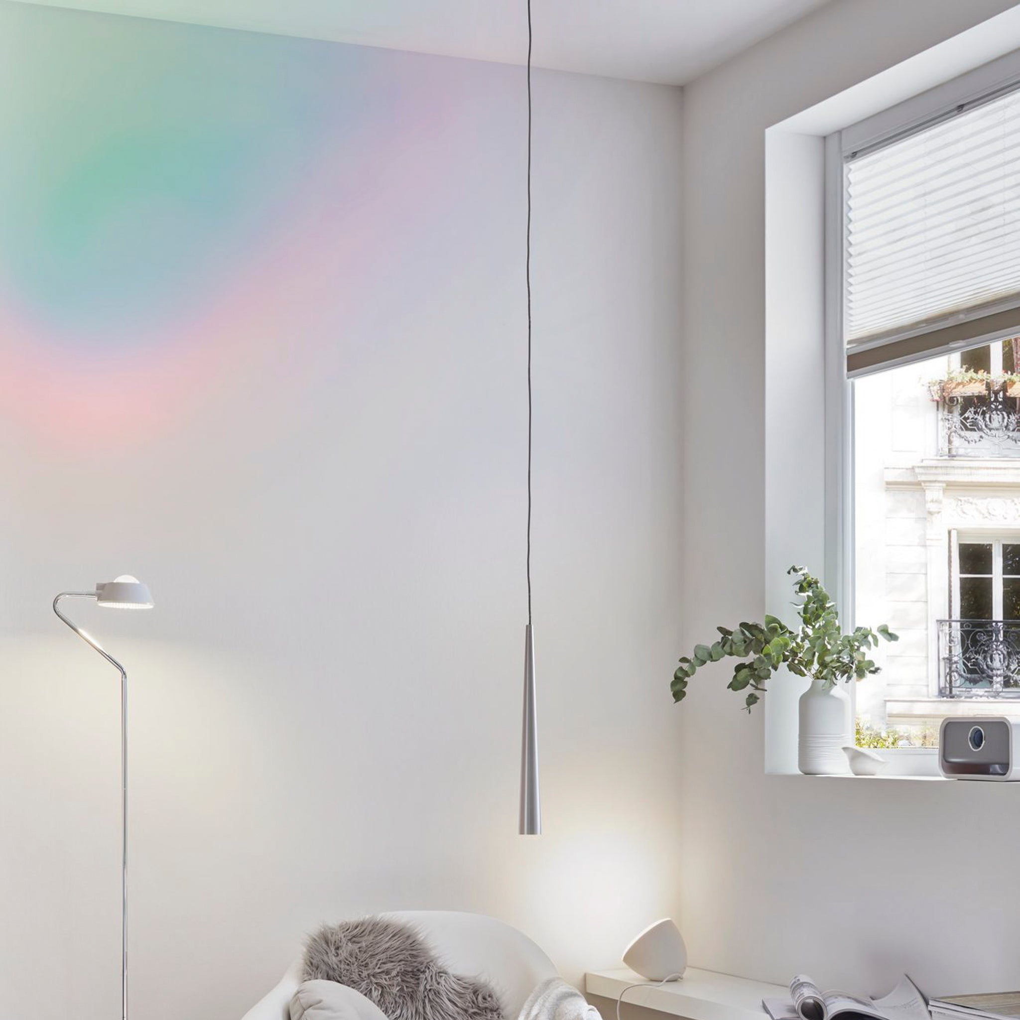 A minimalist room highlights the Paulmann Gutta 8.5W LED Pendant Light in Brushed Aluminium, with its adjustable cable casting a warm glow next to a floor lamp and window. Soft pastel shades complement the white walls, while a plant sits gracefully on the windowsill.