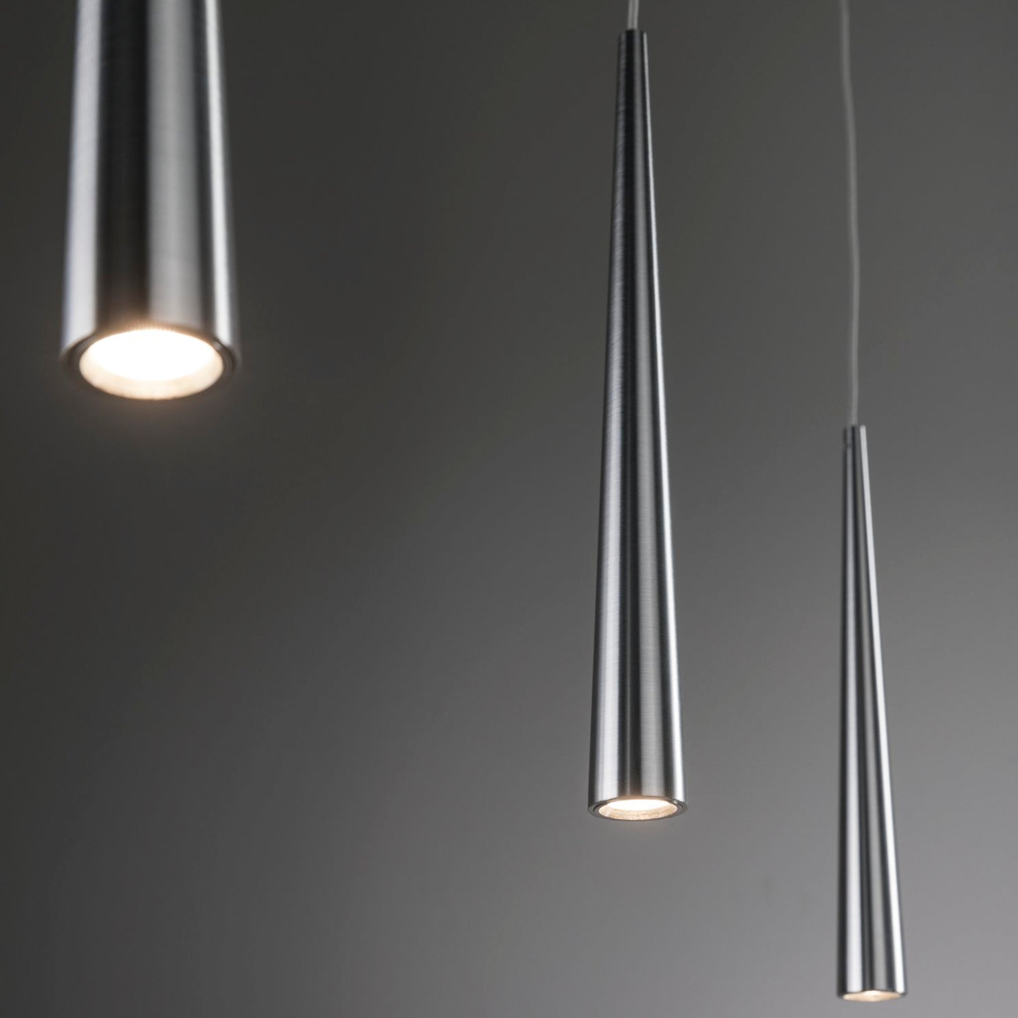 The Paulmann - Gutta 23W LED Dimmable 3 Lamp Pendant Light in Brushed Aluminium, featuring sleek, metallic cone-shaped designs and suspended by adjustable cables, hangs elegantly against a dark grey background.