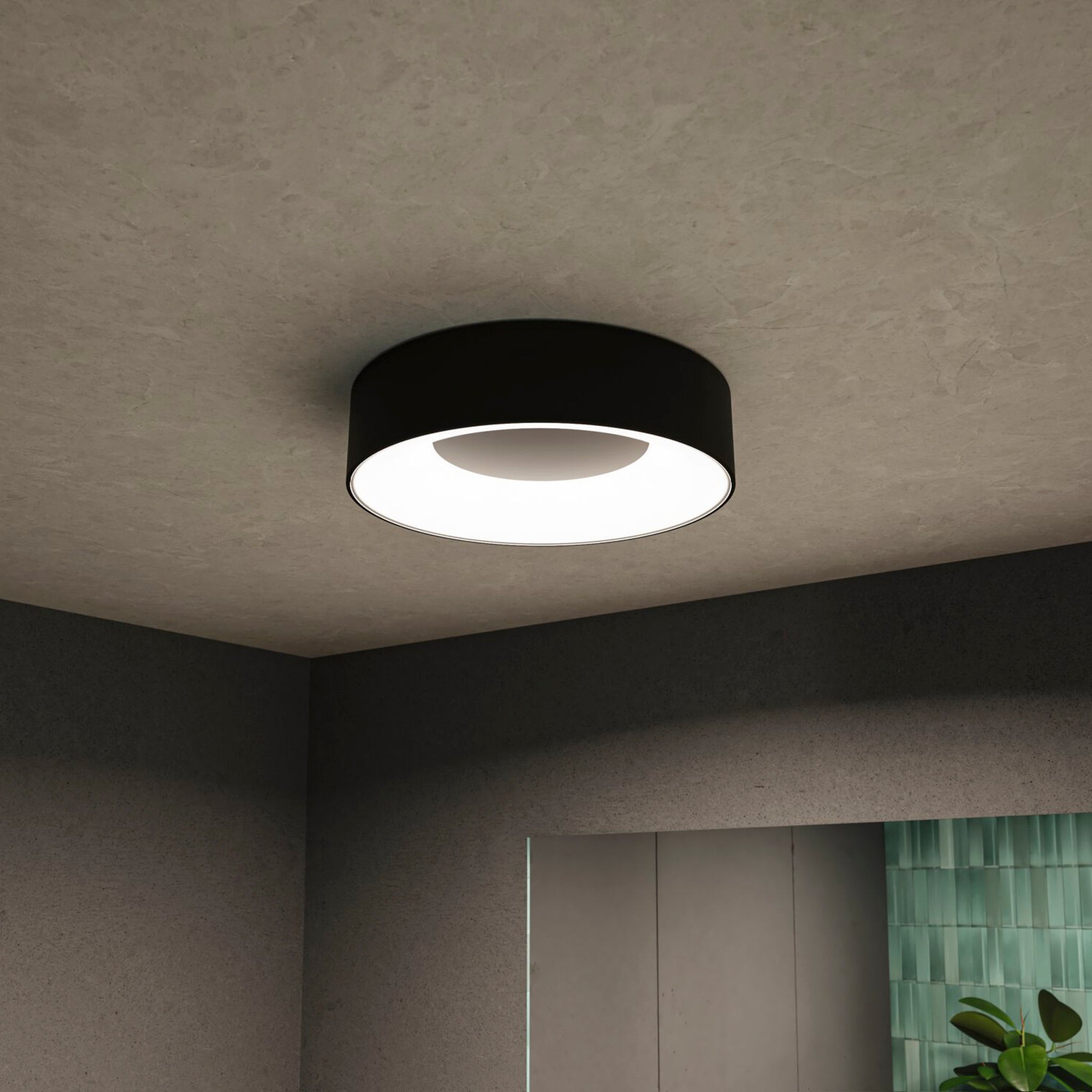 The Paulmann - Casca 18W LED Switch 1500lm Ceiling Light in Black Matt elegantly illuminates a dimly lit room with gray walls, providing a subtle lighting effect that is complemented by a touch of greenery.