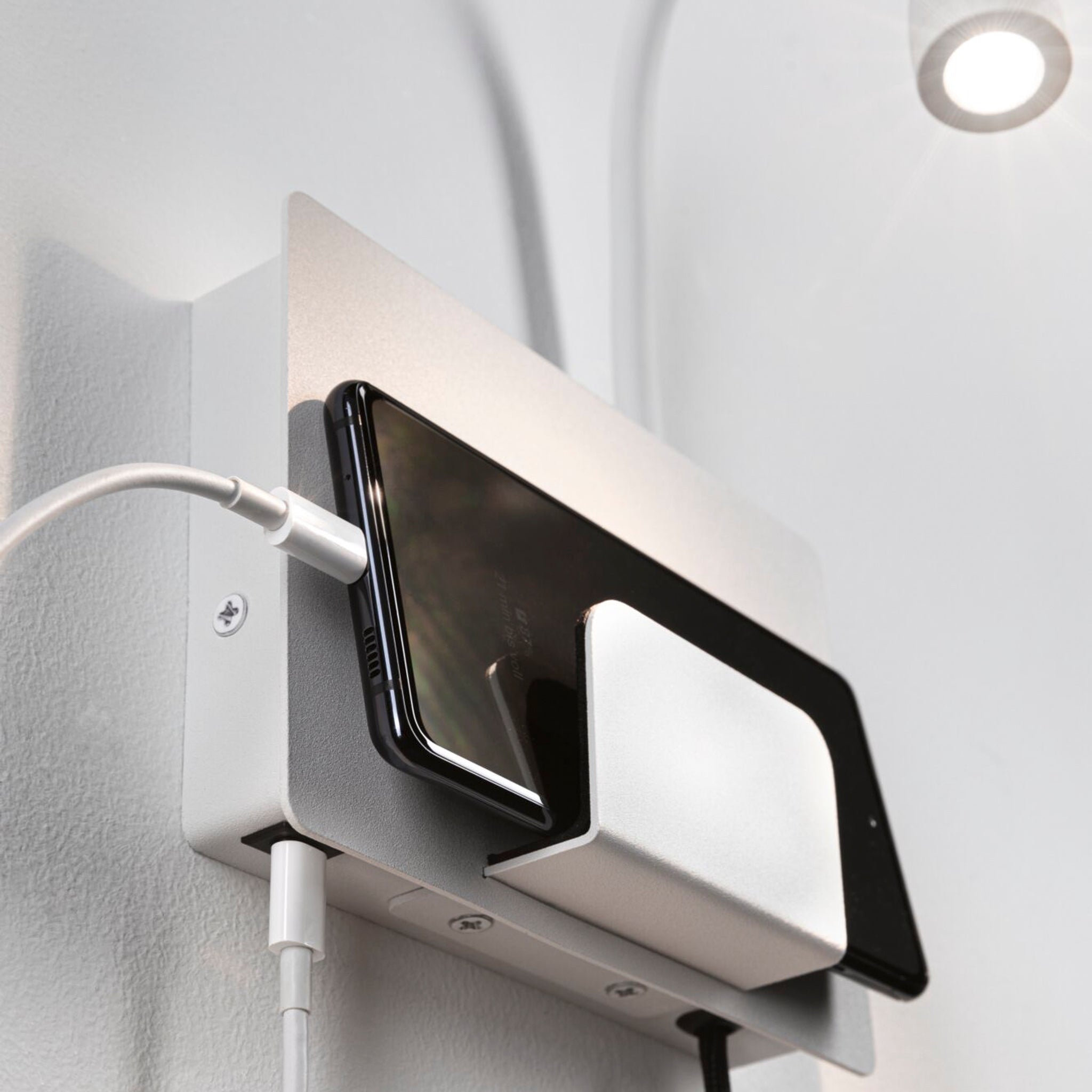 The Paulmann Halina 3.5W LED Wall Light in white matt charges a smartphone on a wall-mounted holder under an LED-lit spotlight, connected via the USB charging port.