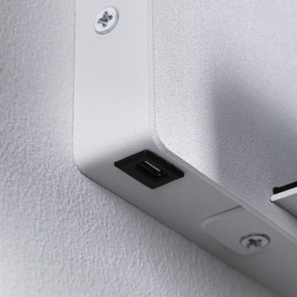 Close-up of a corner of a white matt electronic device mounted on a wall, featuring visible screws and a USB port for USB Type-C charging, seamlessly integrating with the Paulmann - Halina 3.5W LED Wall Light's state-of-the-art illumination.