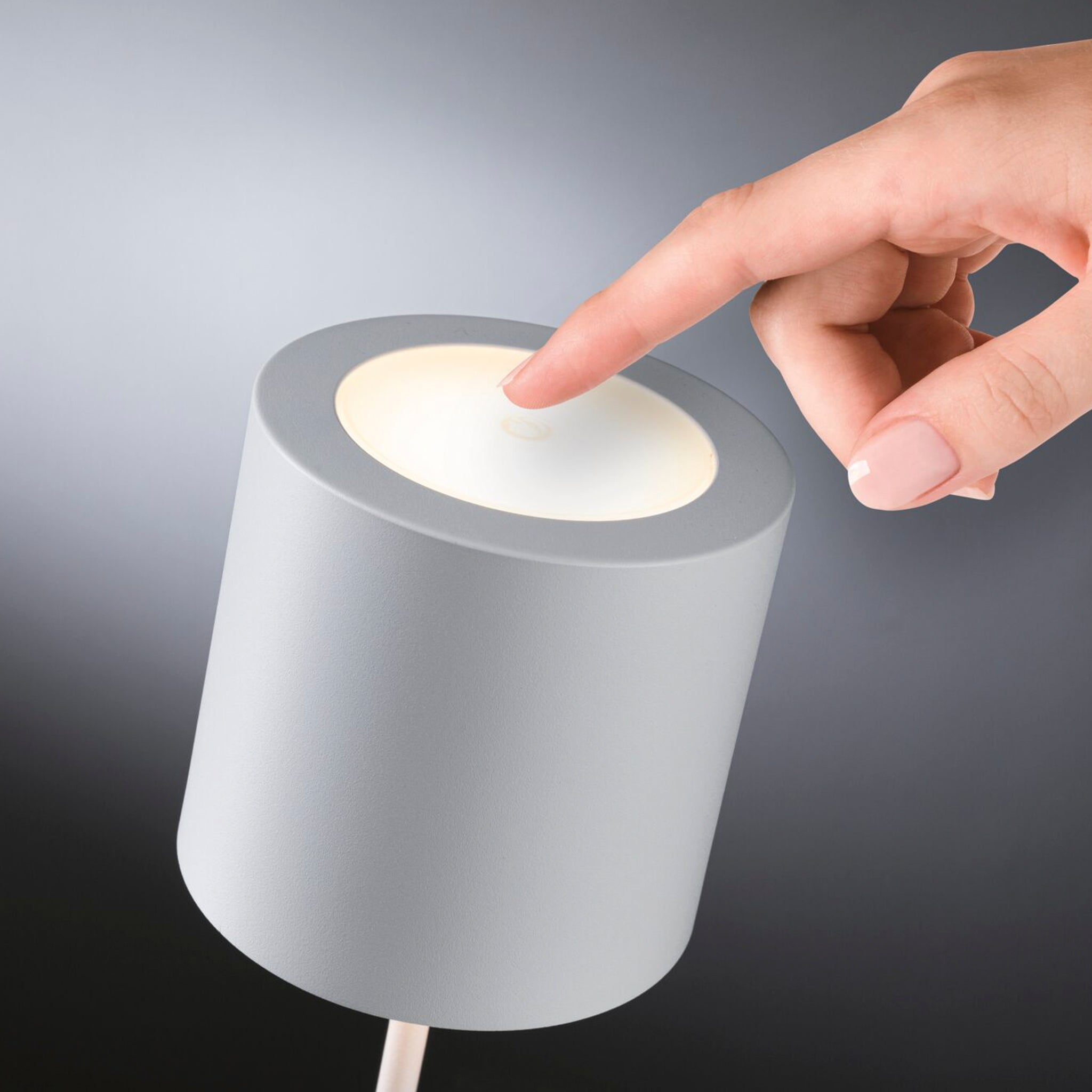 A hand elegantly engages the touch dimming feature on the sleek, white matte cylindrical Paulmann Tuni 2.8W LED Mobile Table Lamp to illuminate its gentle glow. This battery-powered table luminaire combines elegance and portability, making it a versatile addition to any space.