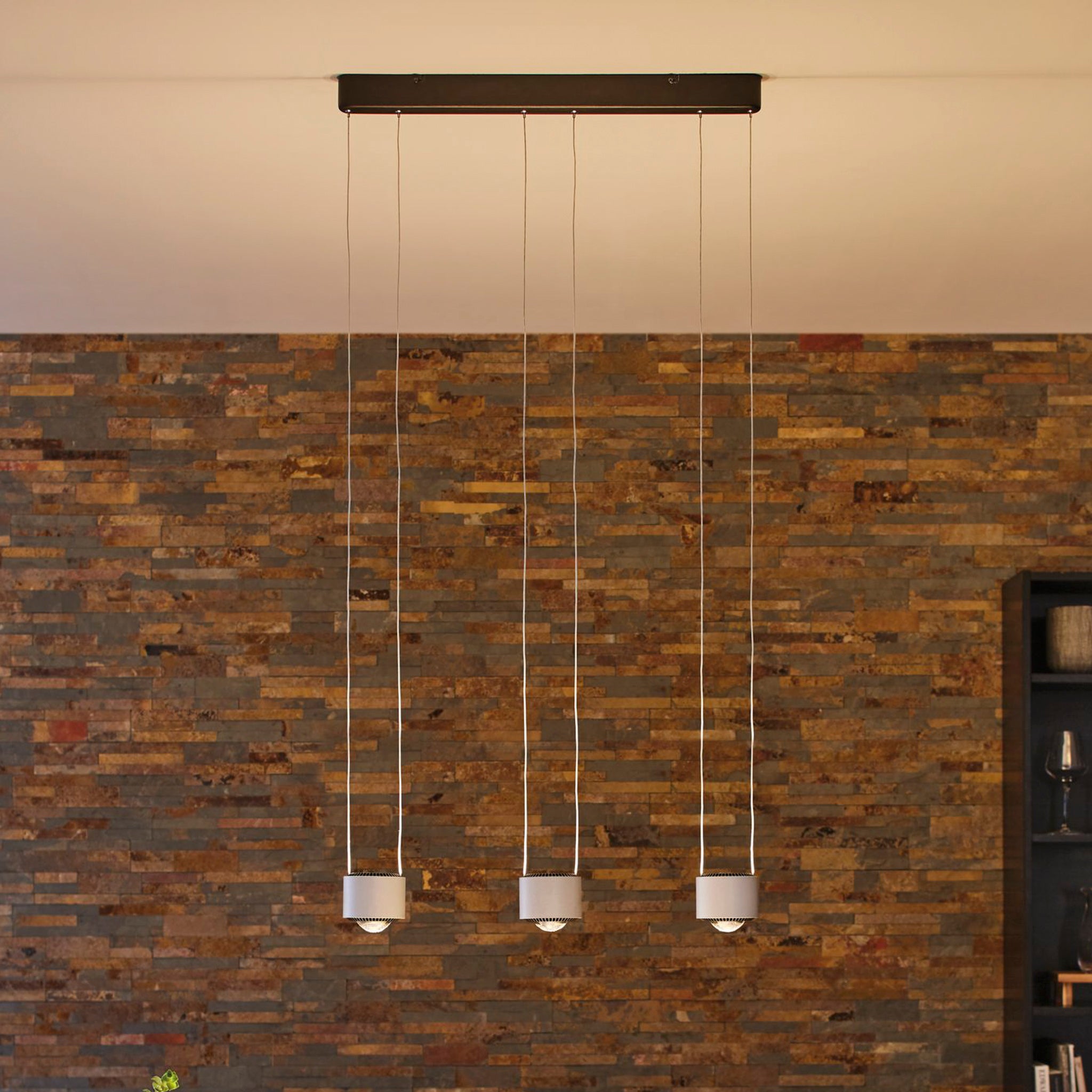 The Paulmann - Aldan 28W LED Dimmable 3 Lamp Pendant Light in brushed aluminium elegantly hangs in front of a rustic brick wall, with its adjustable pendant lengths perfectly enhancing the modern aesthetic.