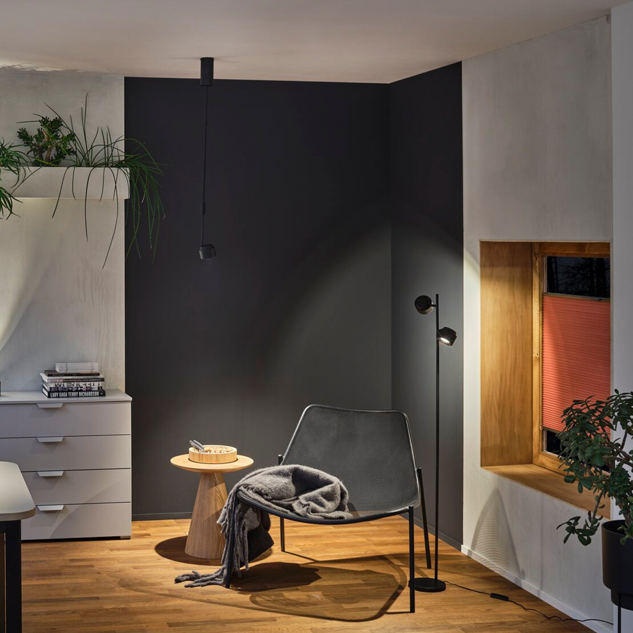 A modern room with a black accent wall, chair, side table, plants, dresser, and window seat adorned with a red blind is enhanced by the Paulmann - Puric Pane 6W LED Dimmable Smart Home Zigbee 3.0 Pendant Light in Black for flexible lighting design.