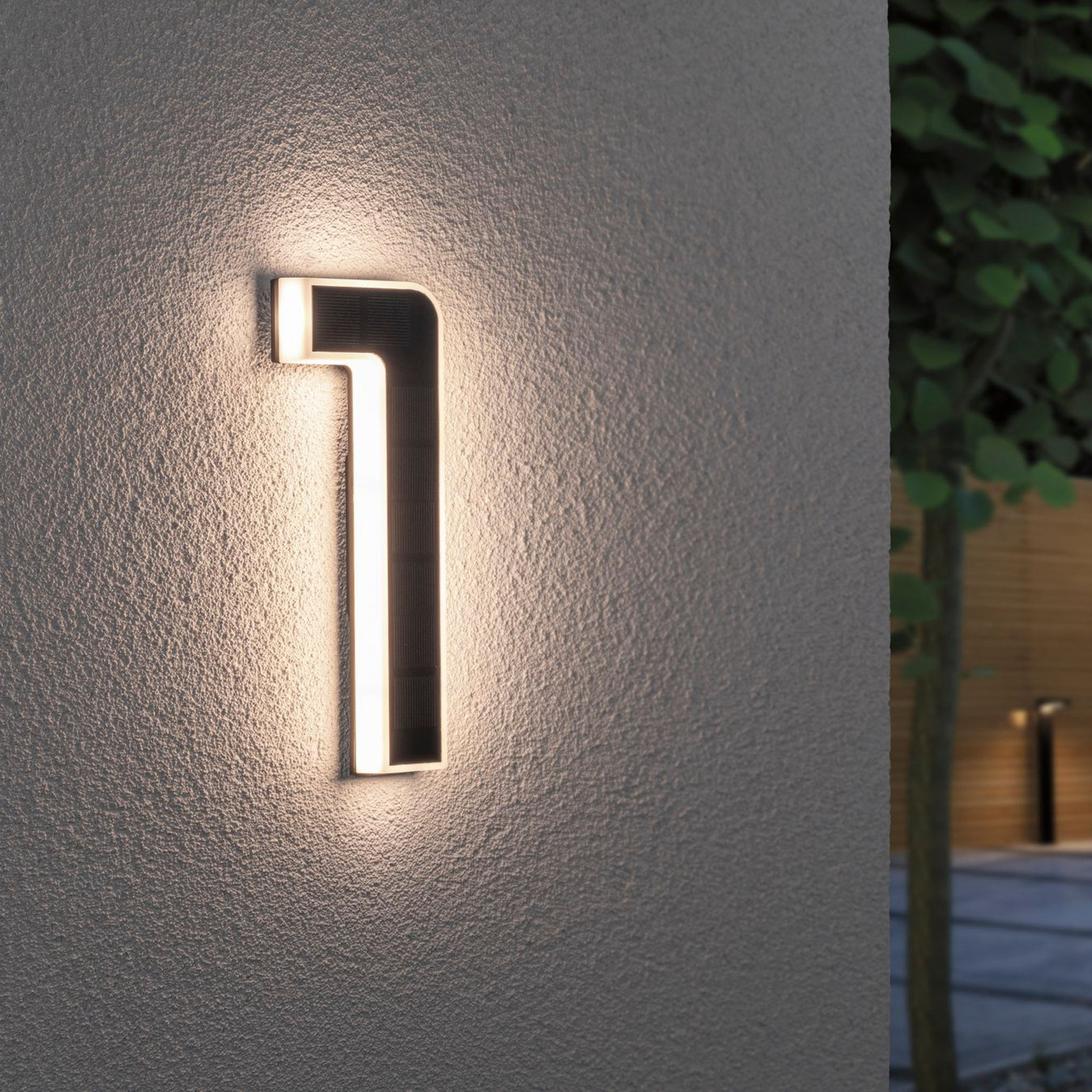 The Paulmann Outdoor Solar House Number 1, featuring a LED 0.2W light and replaceable battery in black, is elegantly mounted on a textured wall. It emits soft lighting while a blurred tree provides a serene backdrop.
