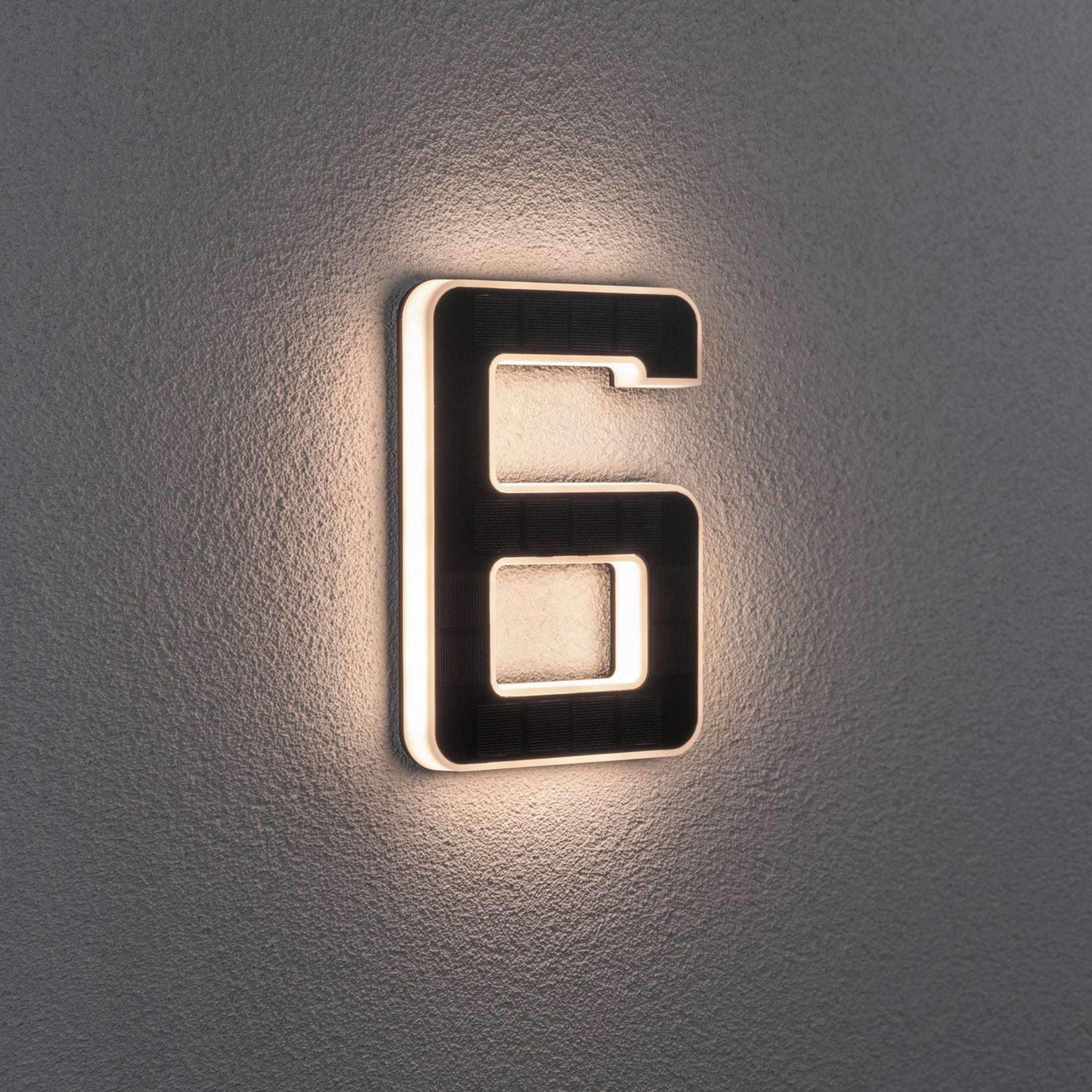 A Paulmann Outdoor Solar House Number 6 LED 0.2W with a replaceable battery in black is mounted on a textured gray wall, casting a soft glow around its edges.