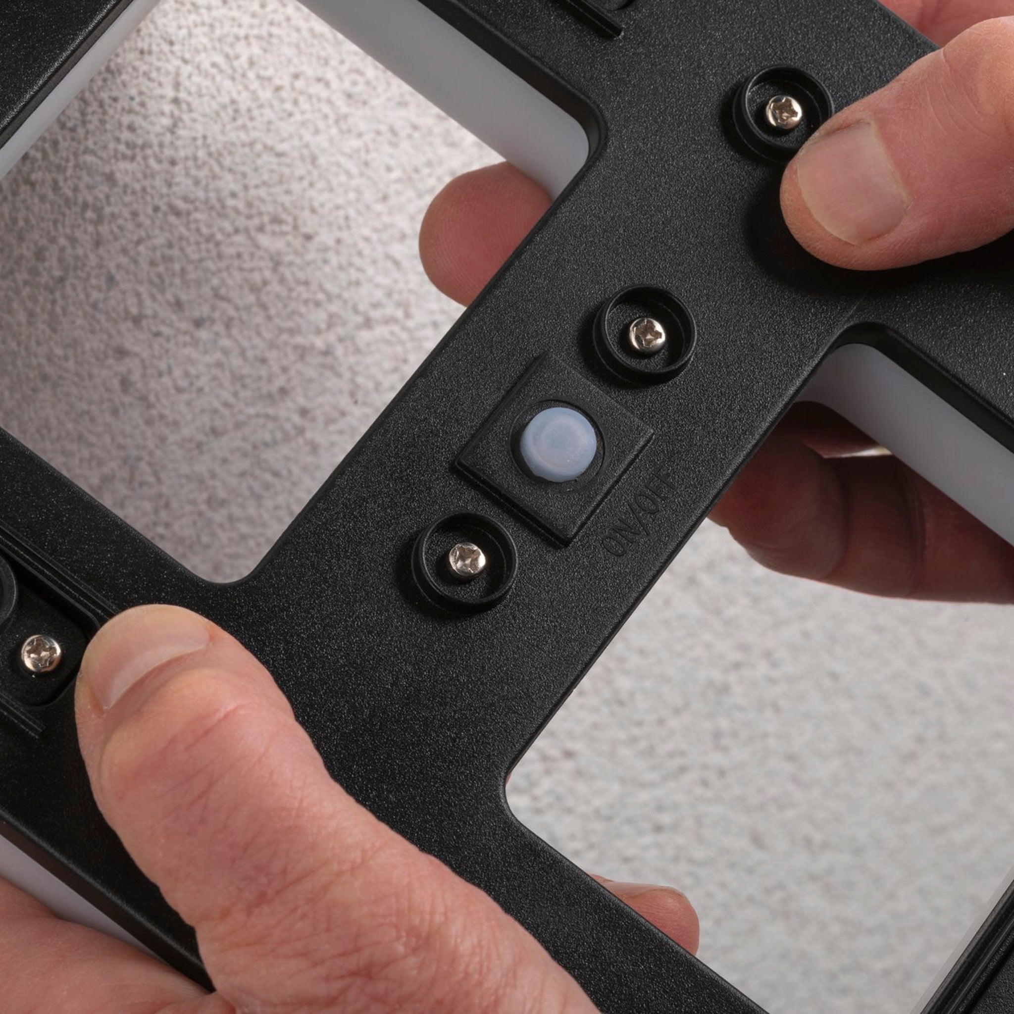 Hands holding a Paulmann Outdoor Solar House Number 7 LED 0.2W with Replaceable Battery in Black, equipped with screws, against a textured white surface, ready for installation to improve visibility.