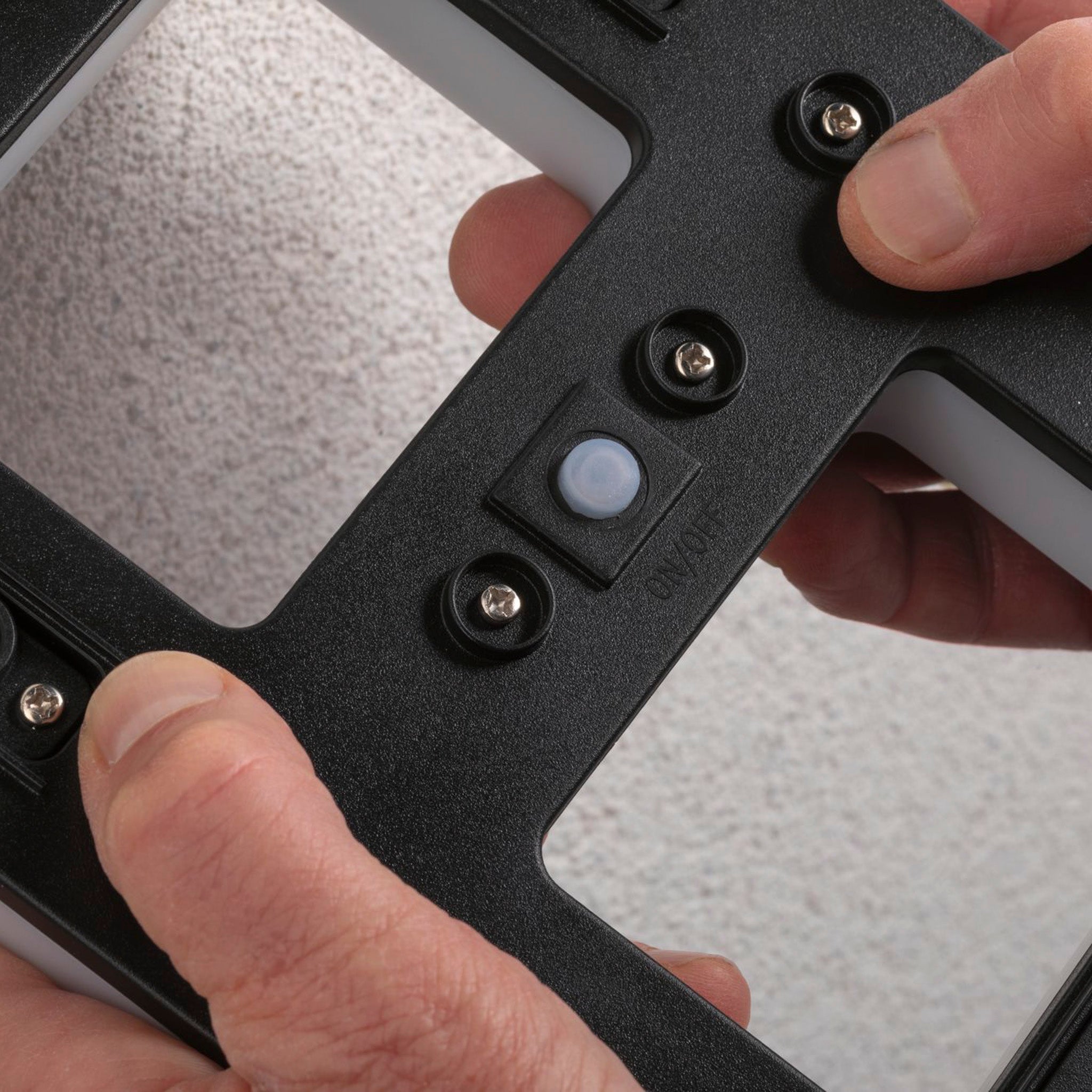 Close-up of hands gripping the black mounting bracket with screws and a small white button designed for the Paulmann Outdoor Solar House Number 8 LED 0.2W with a replaceable battery, set against a gray background.