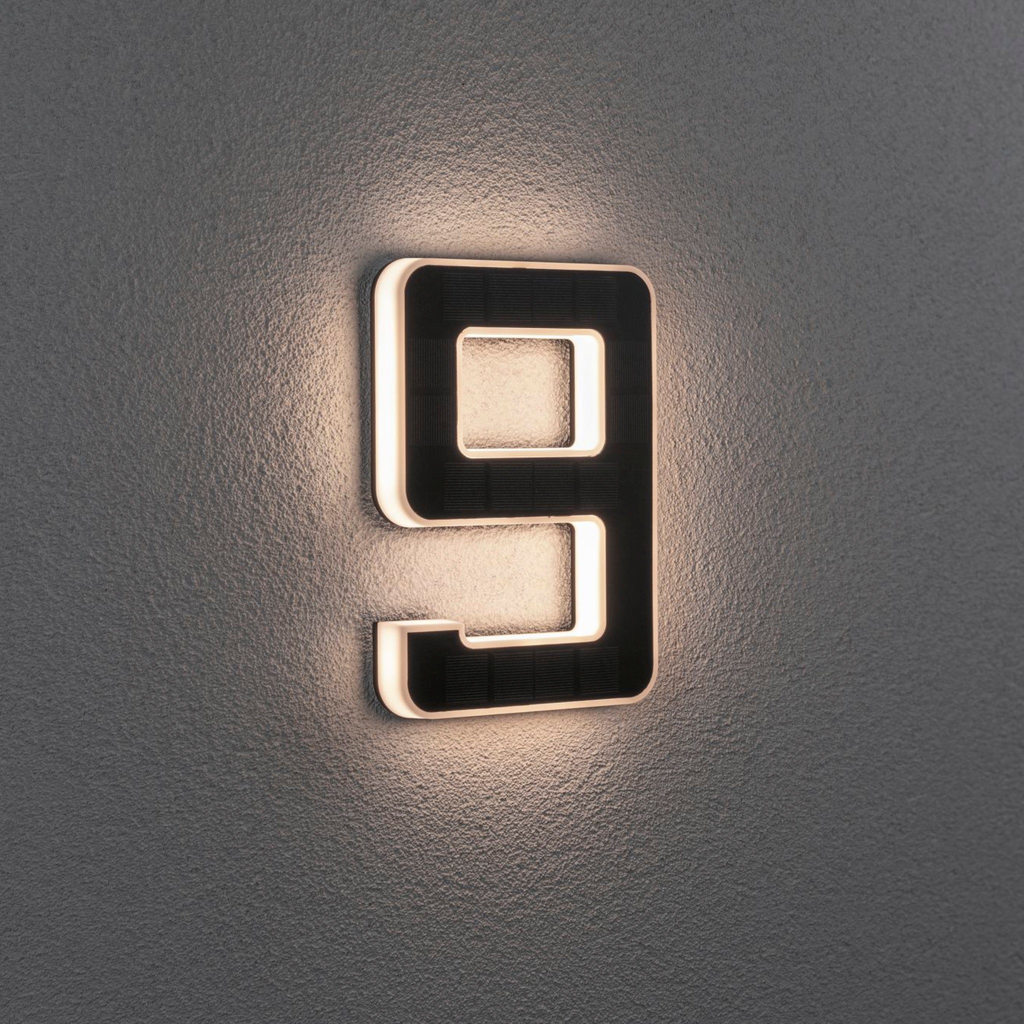 The Paulmann Outdoor Solar House Number 9 LED 0.2W with a replaceable battery in black softly illuminates on the gray wall, functioning as an elegant house number luminaire.