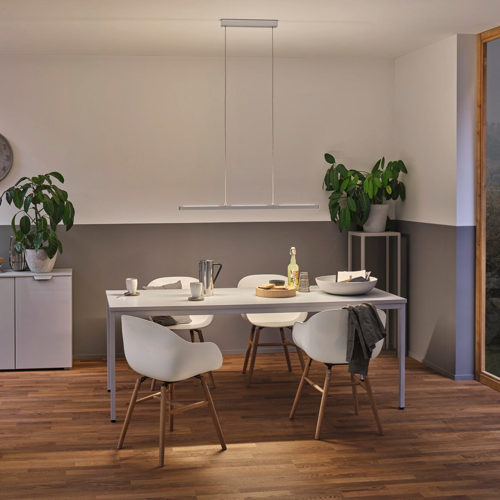 A minimalist dining room featuring a white table, modern chairs, and potted plants is gently illuminated by the Paulmann Lento 37W LED Dimmable Pendant Light in Chrome Matt.
