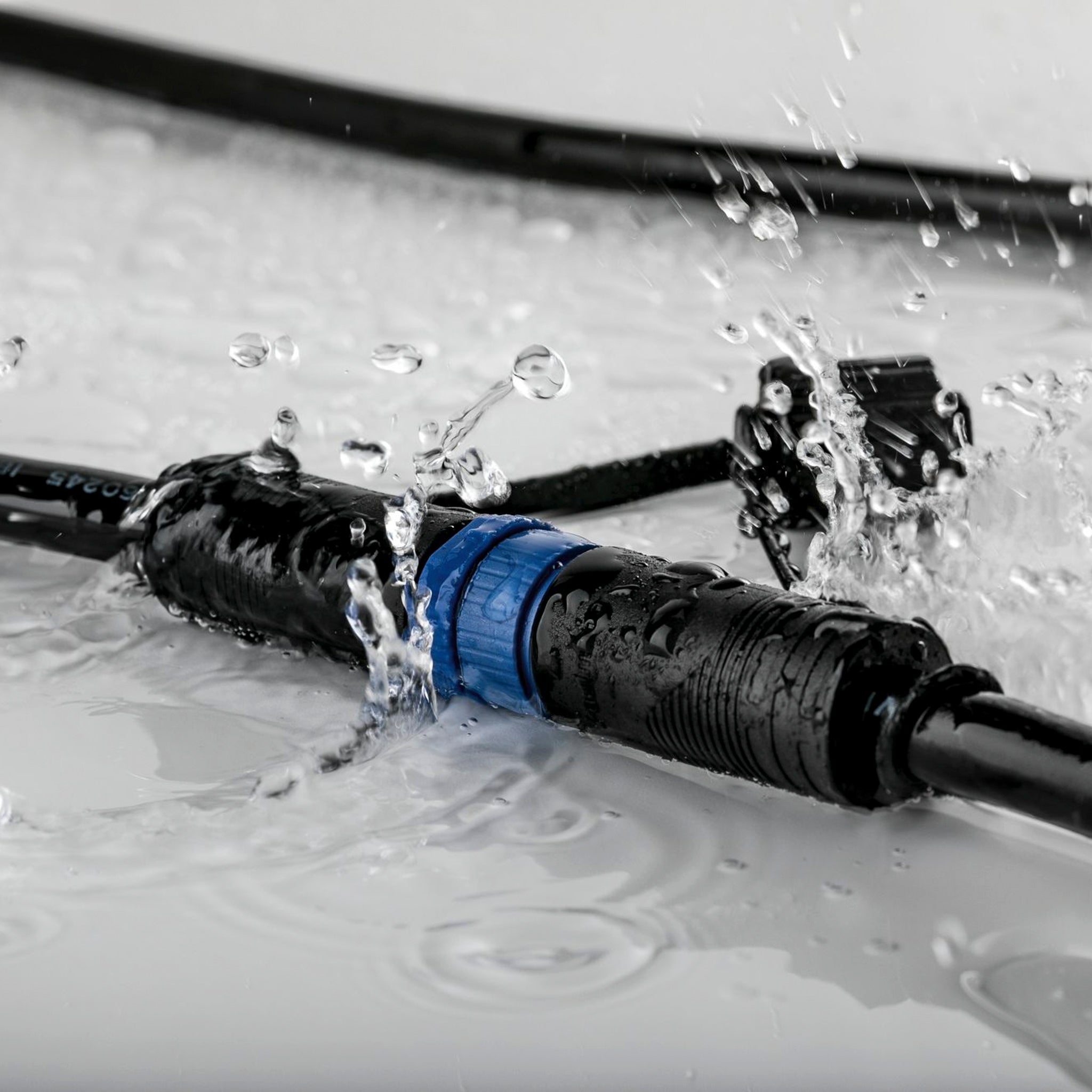 Water splashes around the Paulmann - Plug & Shine Outdoor 10m Cables with 5 Outputs in Black, featuring a blue connector tailored for outdoor use. With IP68 immersion protection, this cable confidently rests on wet surfaces.