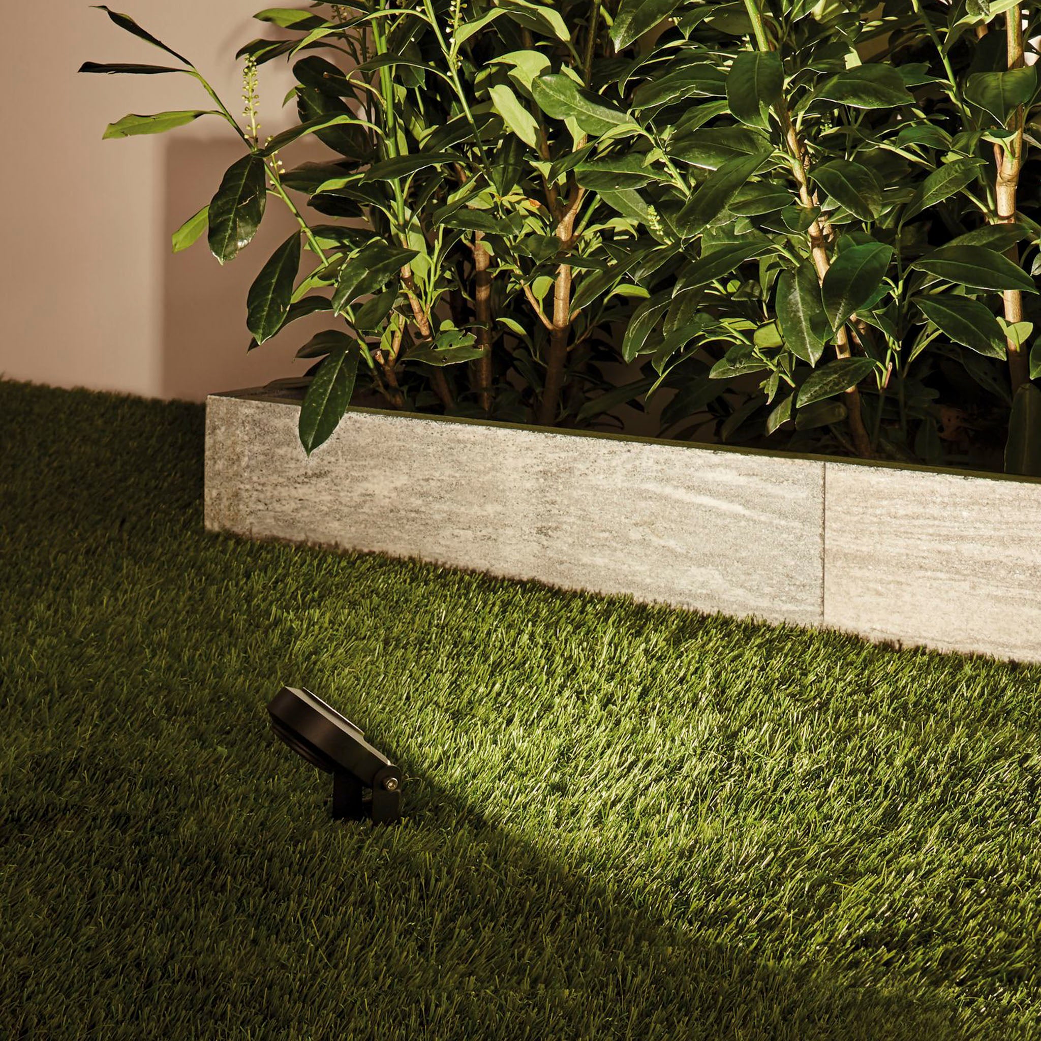 The Paulmann - Plug & Shine Outdoor String 6.3W LED Warm White Individual Spotlight in Anthracite emits a warm white light, lighting up a patch of grass next to a stone planter adorned with tall, leafy plants. Its weatherproof design ensures that this tranquil glow withstands the elements gracefully.