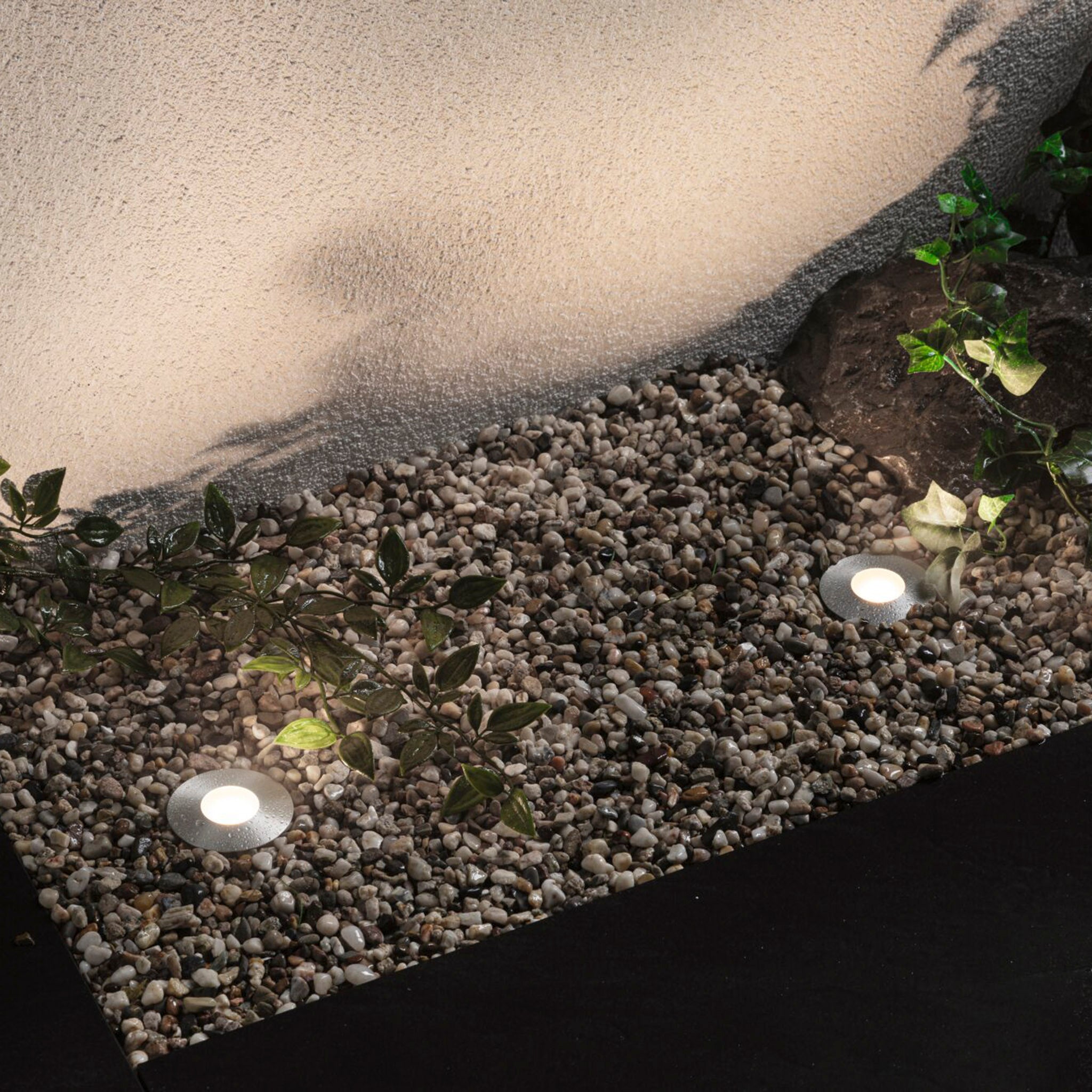 The garden pathway features recessed spotlights and two round ground lights, where leafy plants create shadows on a textured wall, showcasing the elegance of outdoor lighting. It's an ideal setup for those who appreciate the seamless ambiance provided by the Plug & Shine system, including the Paulmann - Plug & Shine Outdoor 2.5W LED Floor Mini Individual Spotlight in Silver.