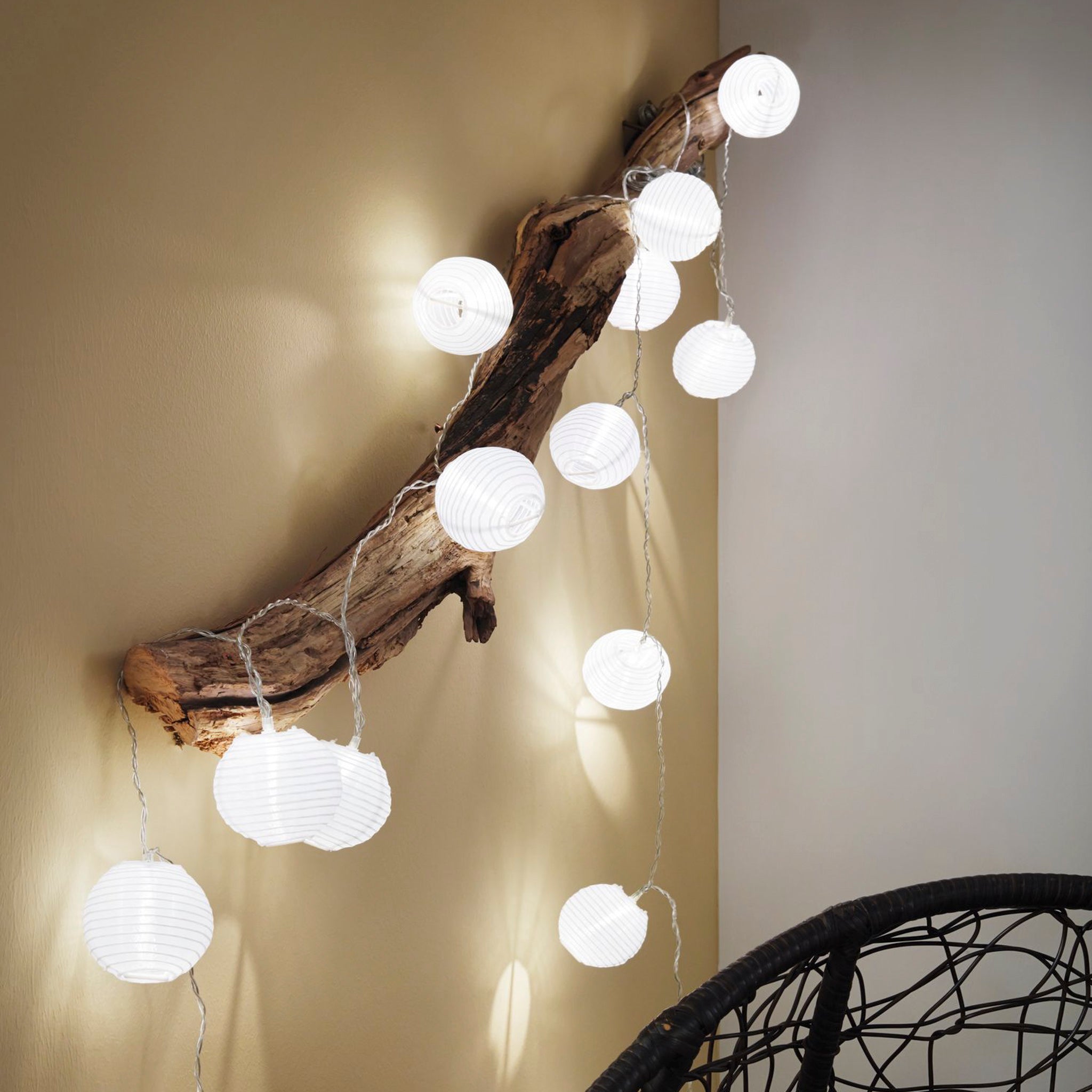 Displaying the Paulmann Outdoor Lampion LED 3.6m Fairy Lights in Warm White on a rustic wooden branch against a beige wall establishes an enchanting outdoor ambiance, as the USB-rechargeable lanterns cast a gentle glow over the partially visible wicker chair below.